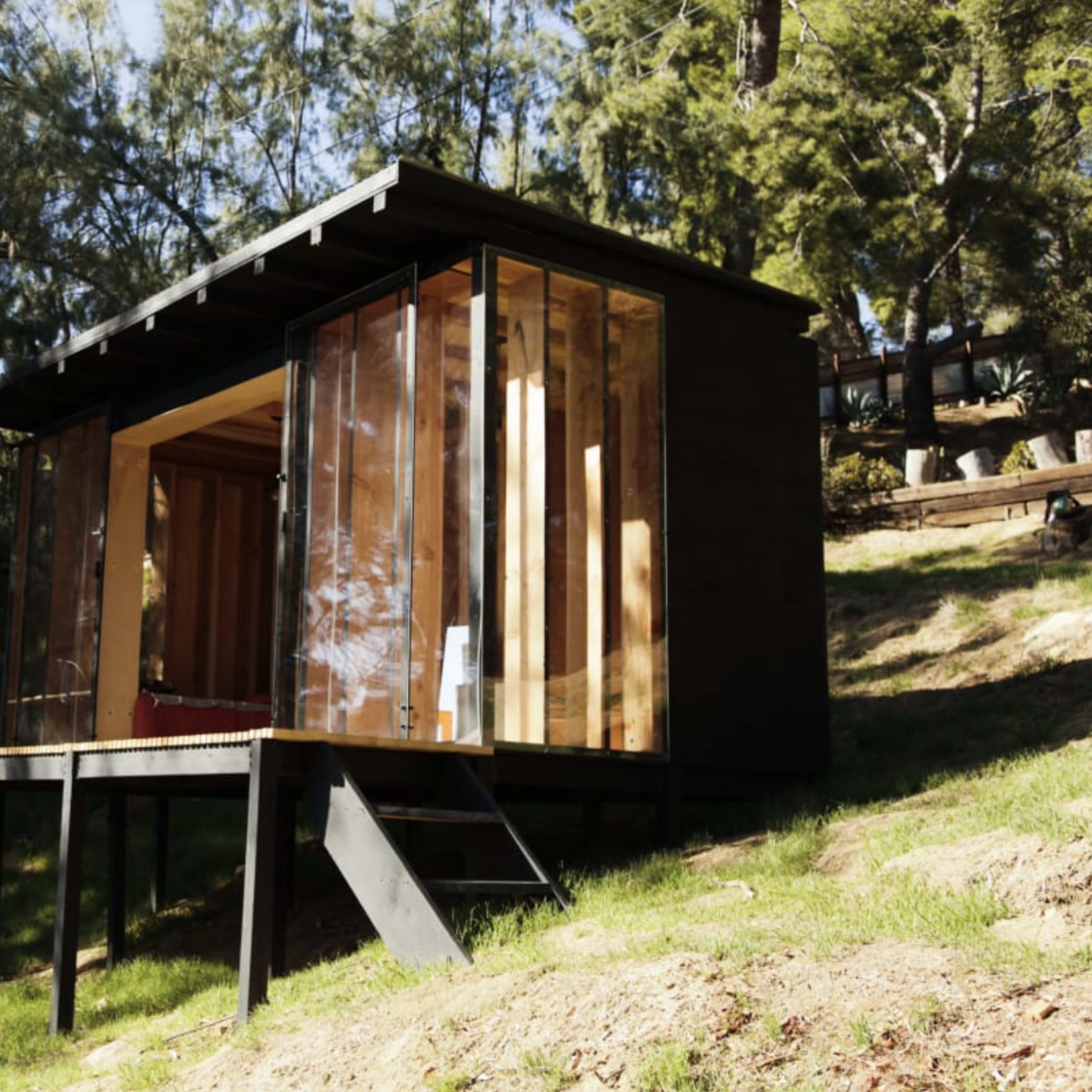A small modern cabin with large glass panels is situated on a grassy slope surrounded by trees.