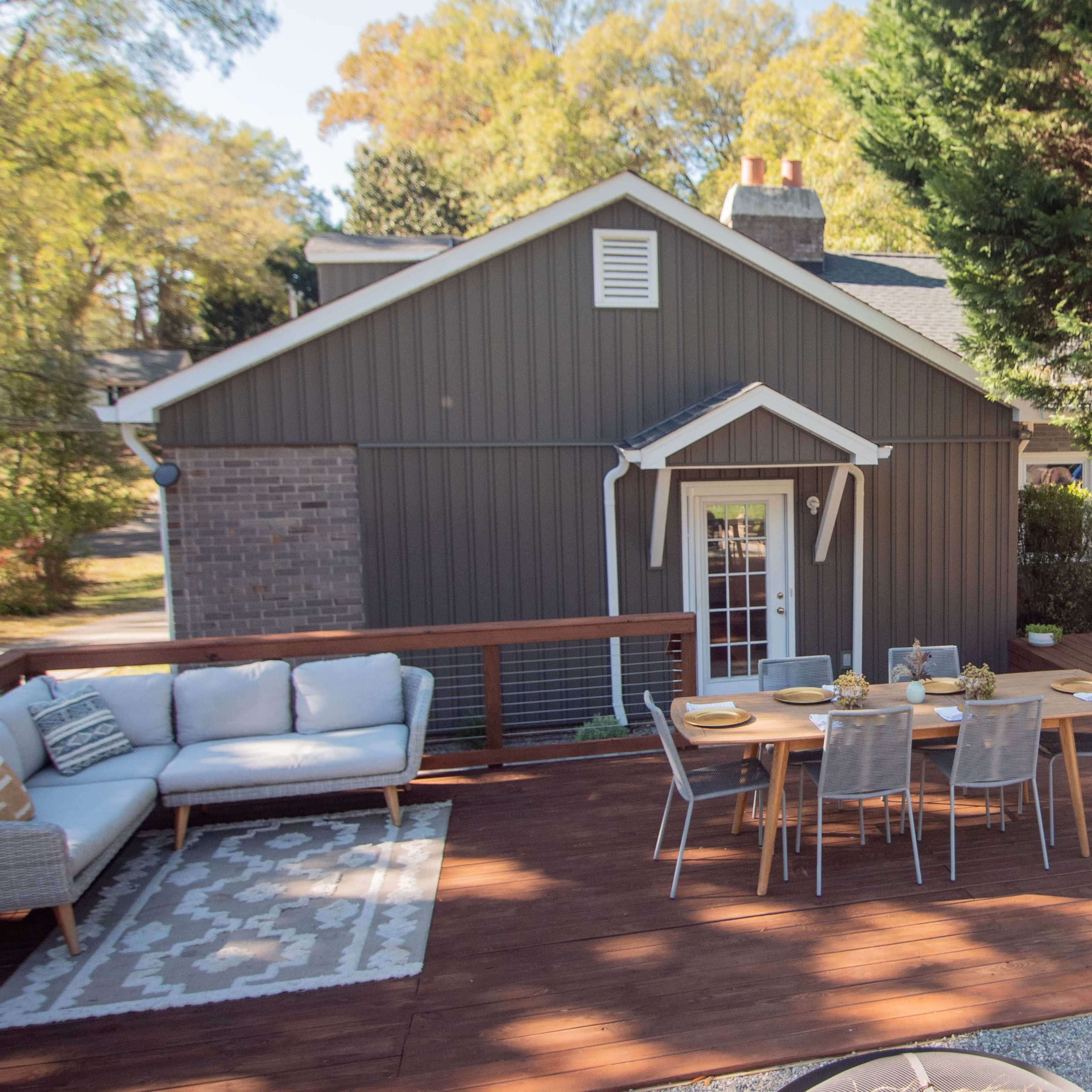 A wooden deck with a dining table and seating area is adjacent to a gray house surrounded by greenery.