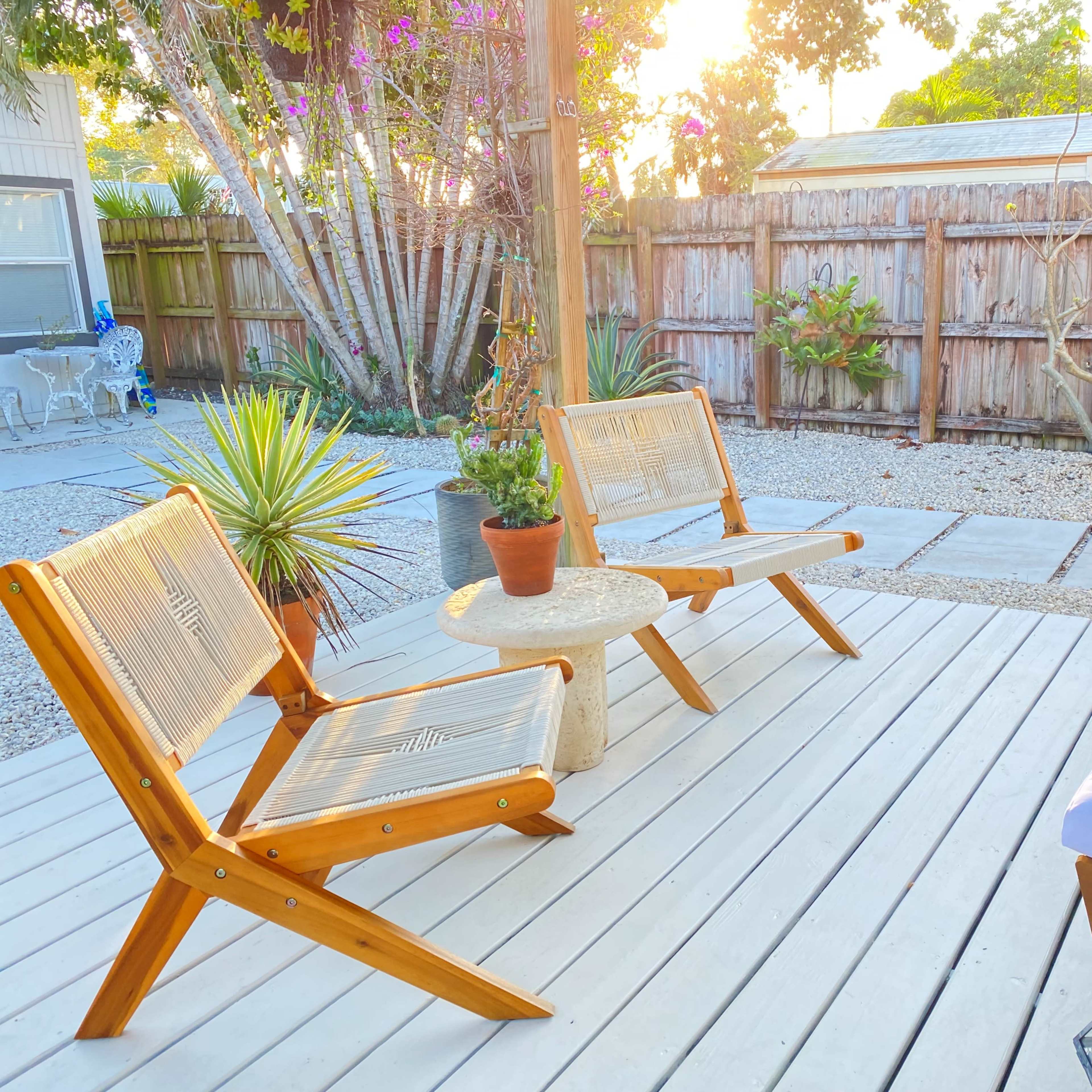 A wooden patio features two chairs and a small round table, surrounded by gravel and tropical plants, with a wooden fence in the background.