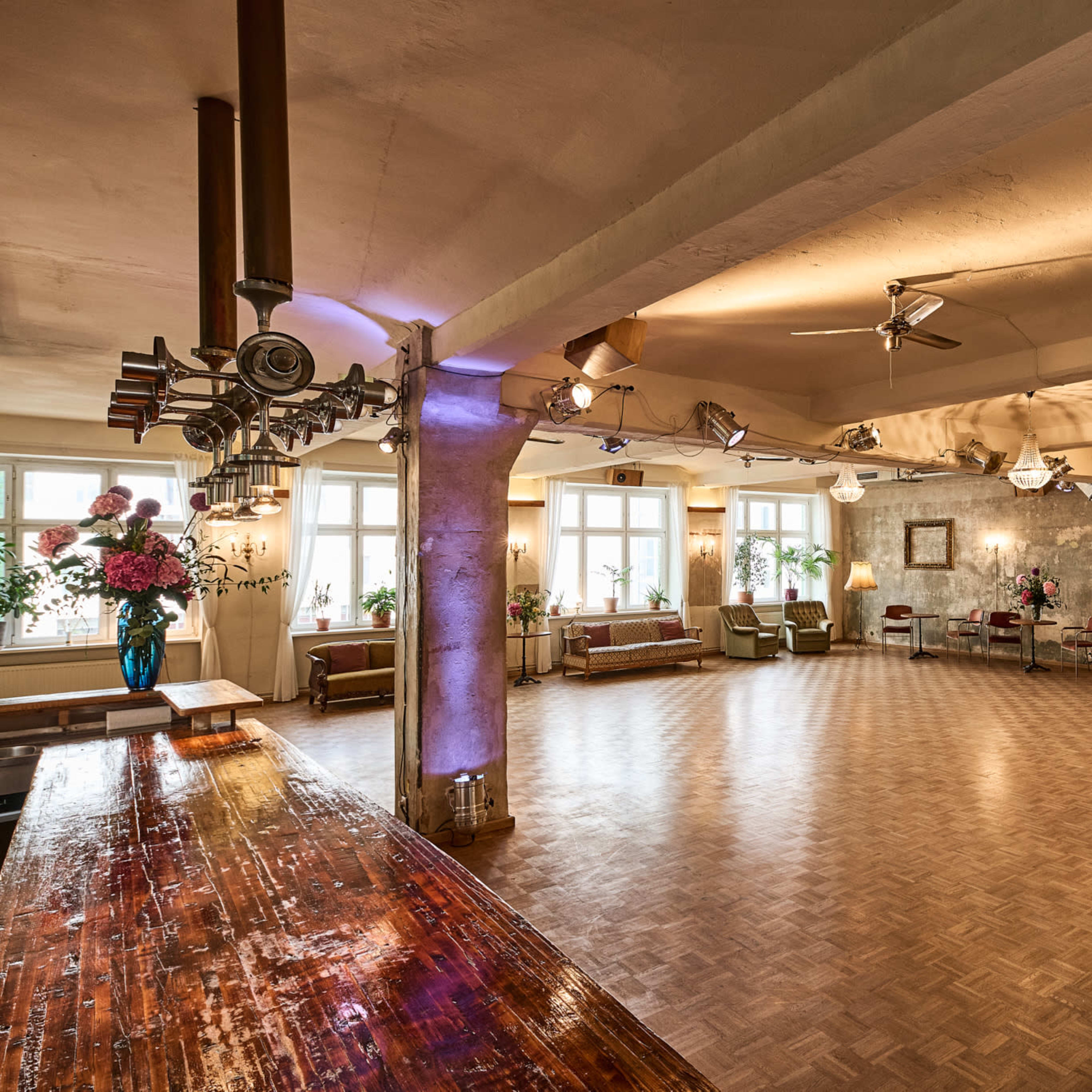 A spacious, well-lit room features wooden floors, a bar area, several seating arrangements, and large windows with plants.