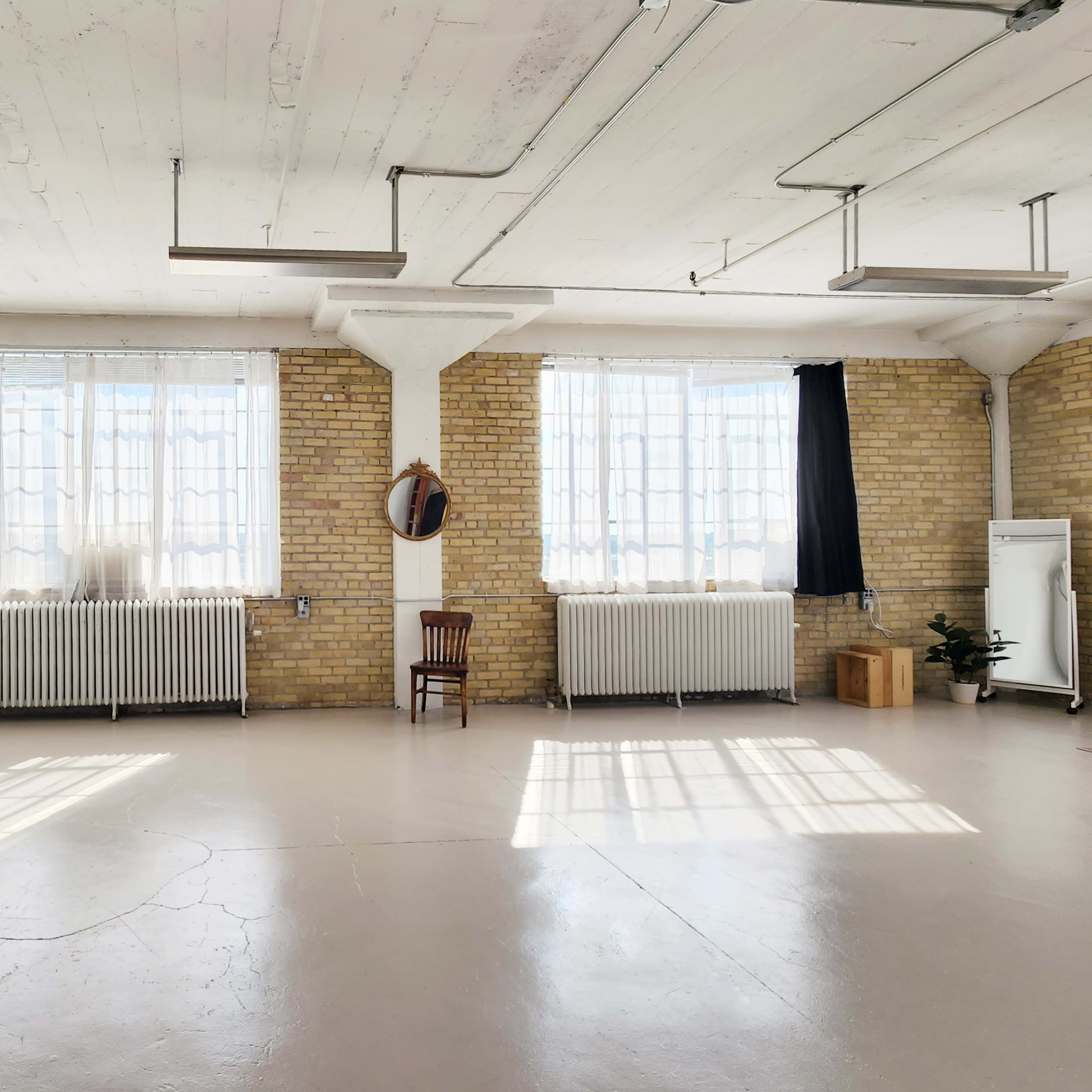A spacious room with exposed brick walls, large windows, and minimal decor features a chair, a mirror, and a potted plant.