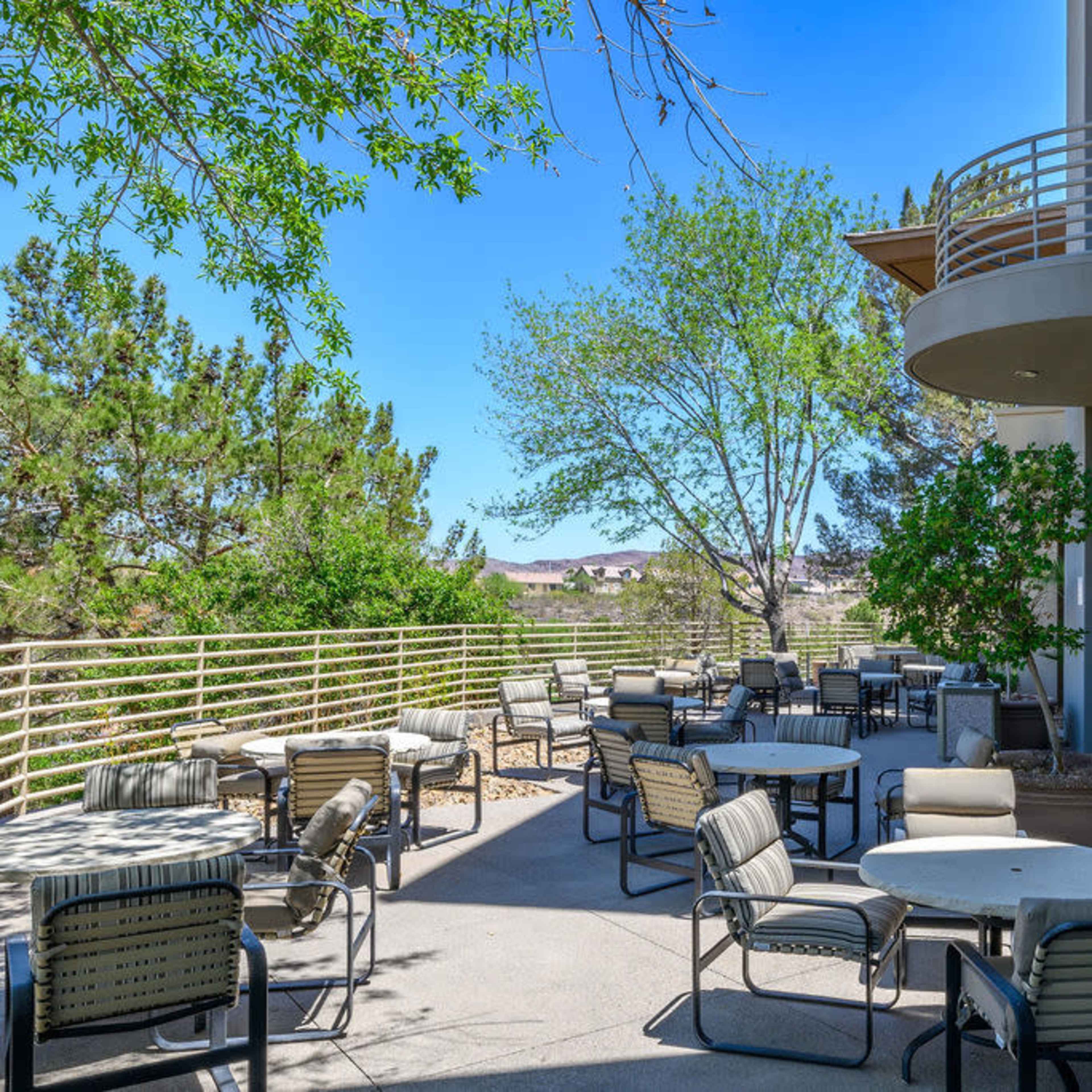 A patio area with multiple tables and chairs is situated next to a building, surrounded by greenery and a clear blue sky.