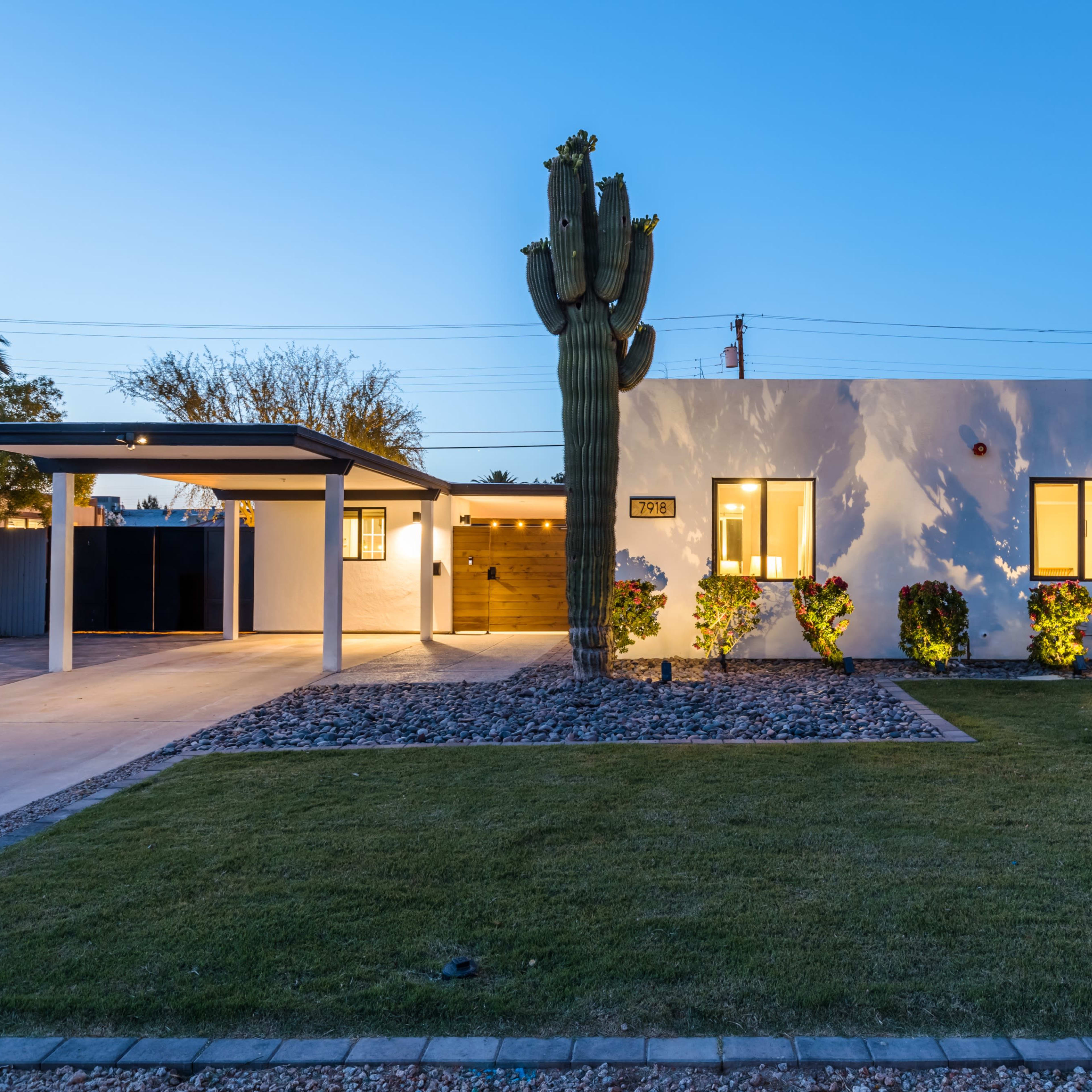 A modern white house with large windows and a carport is situated in a landscaped yard featuring a prominent cactus and gravel accents.
