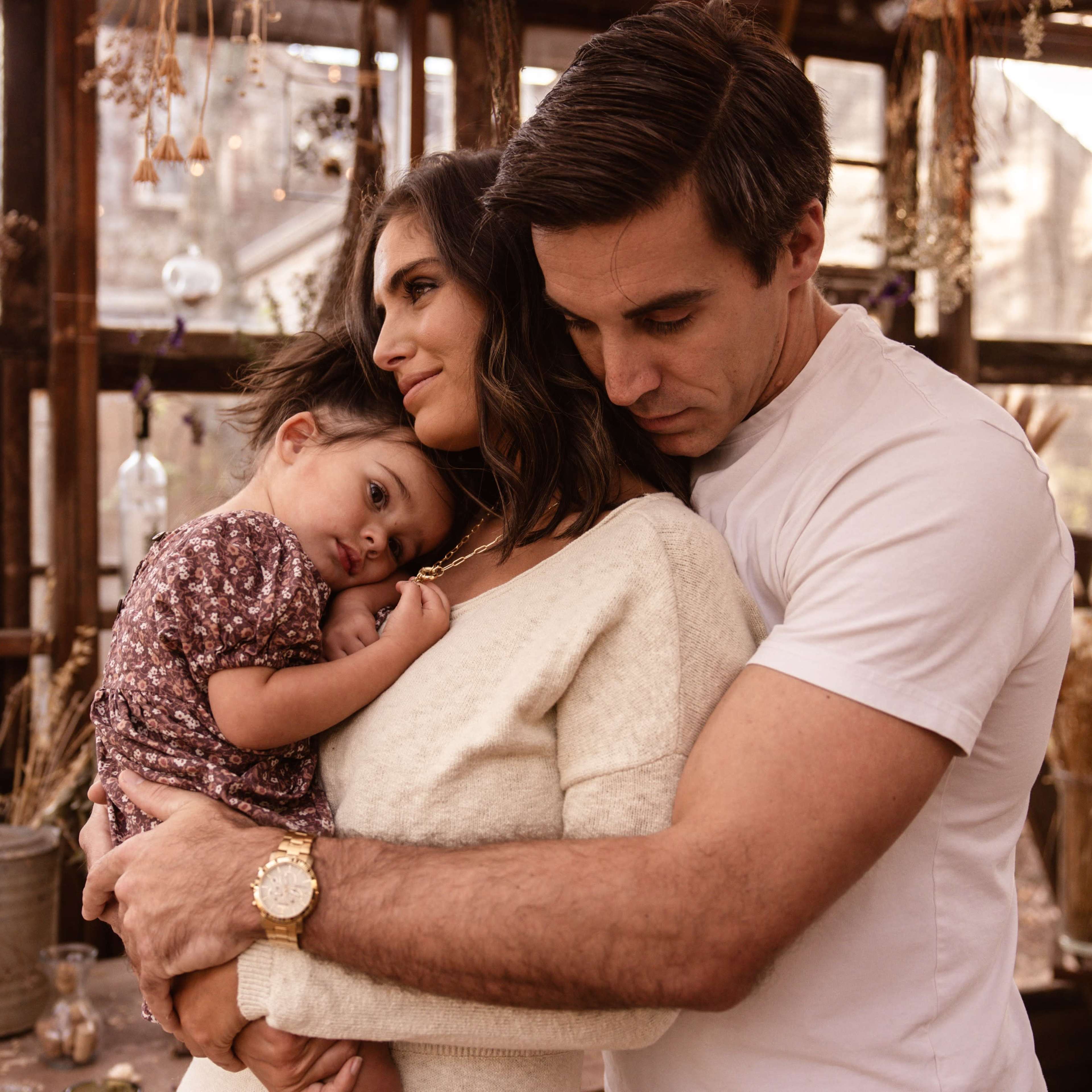 A woman holds a young child close while a man embraces them both in a rustic, plant-filled setting.