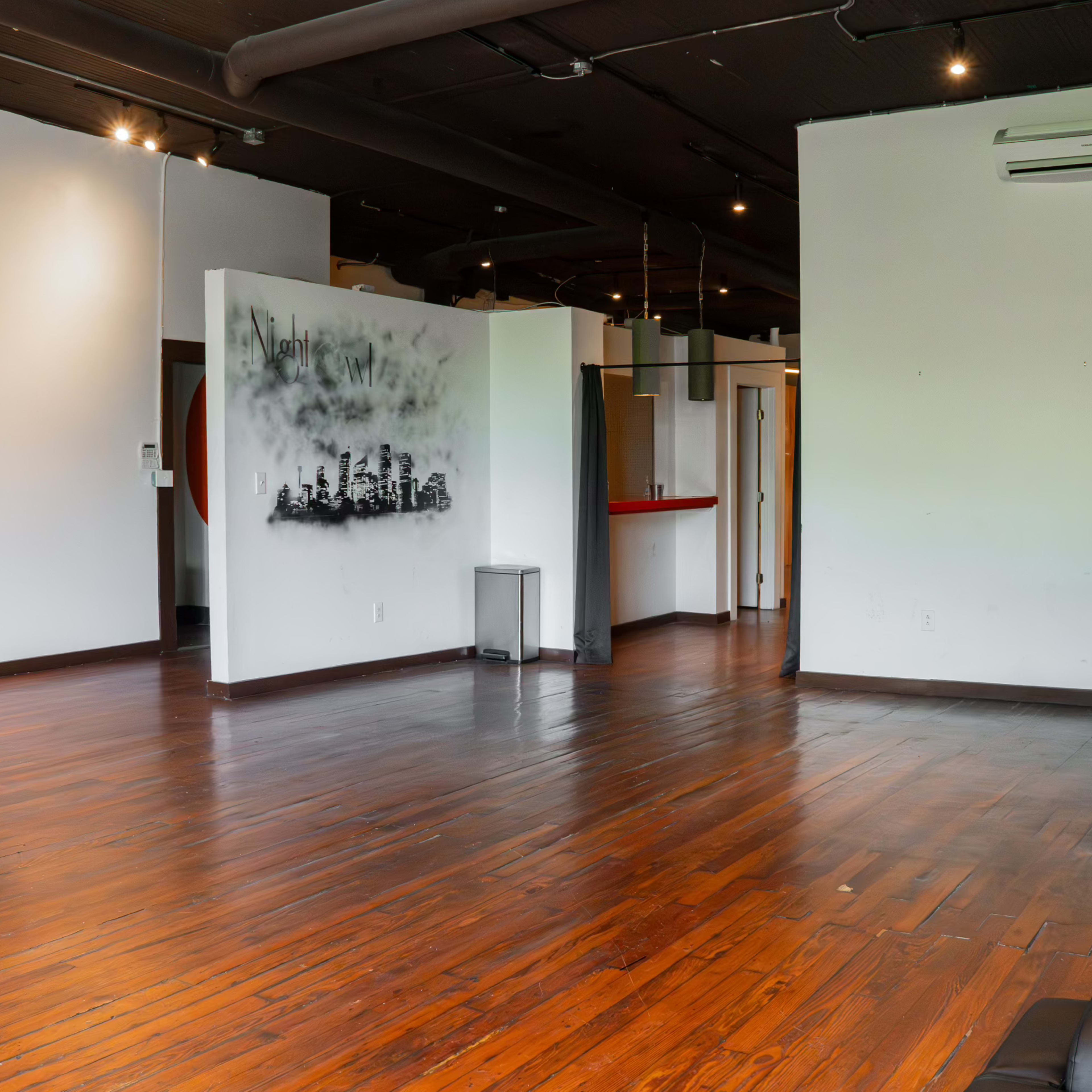 An open room with wooden flooring, a wall featuring a city skyline mural, and several areas including a kitchenette and a small partitioned space.