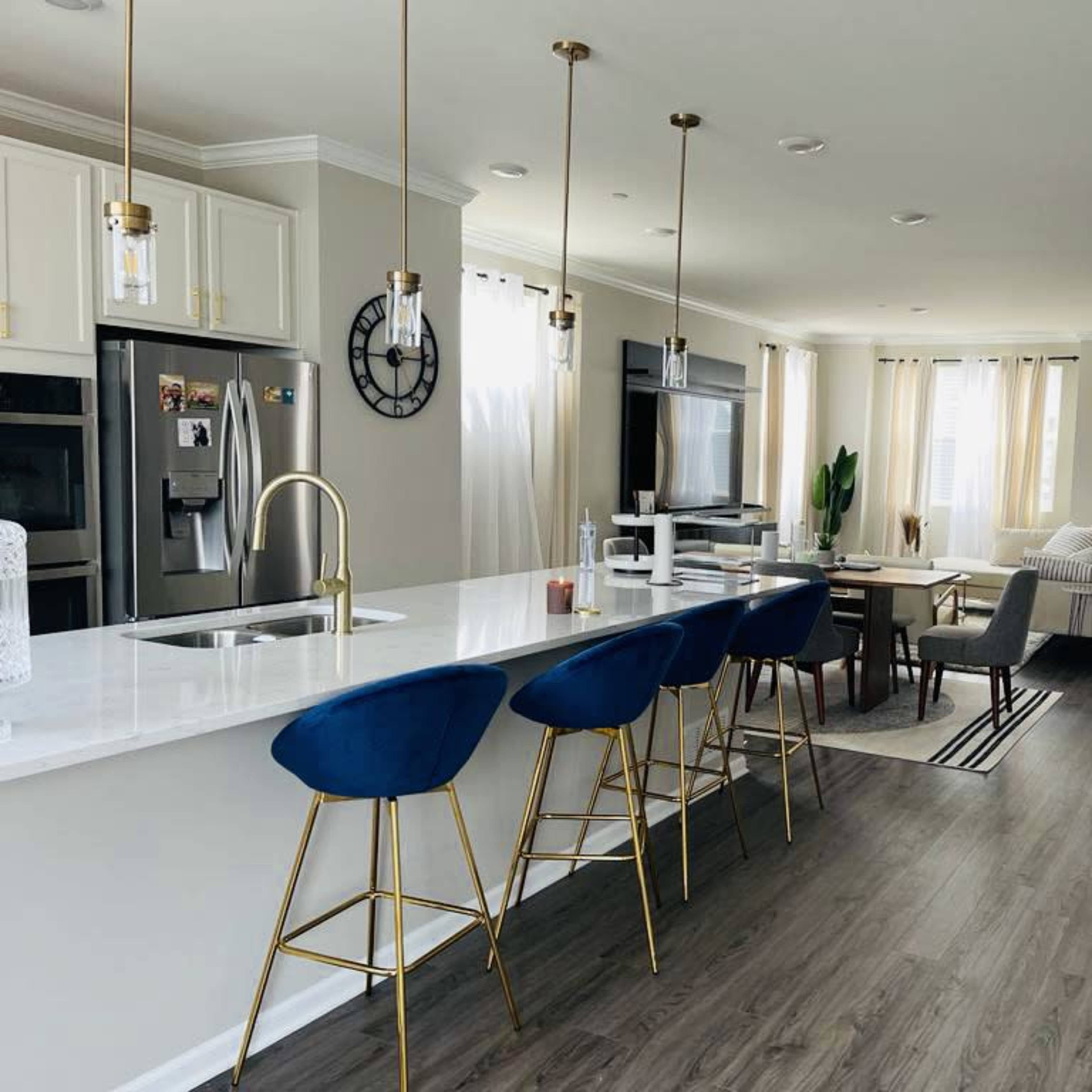 A modern kitchen and living area with a white countertop, blue bar stools, and light-colored furniture.