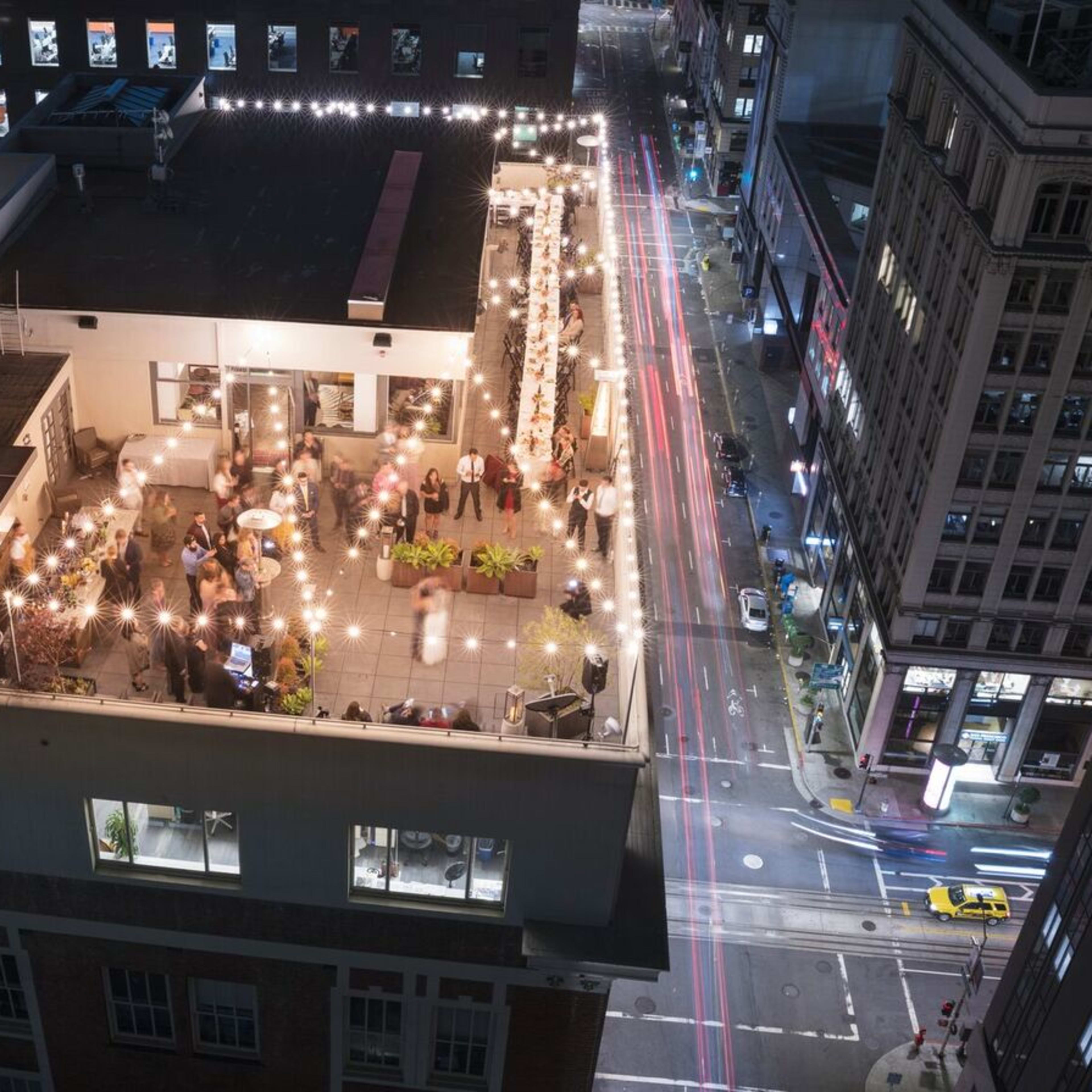 A rooftop terrace with string lights and seating overlooks a city street at night, showcasing a lively gathering.