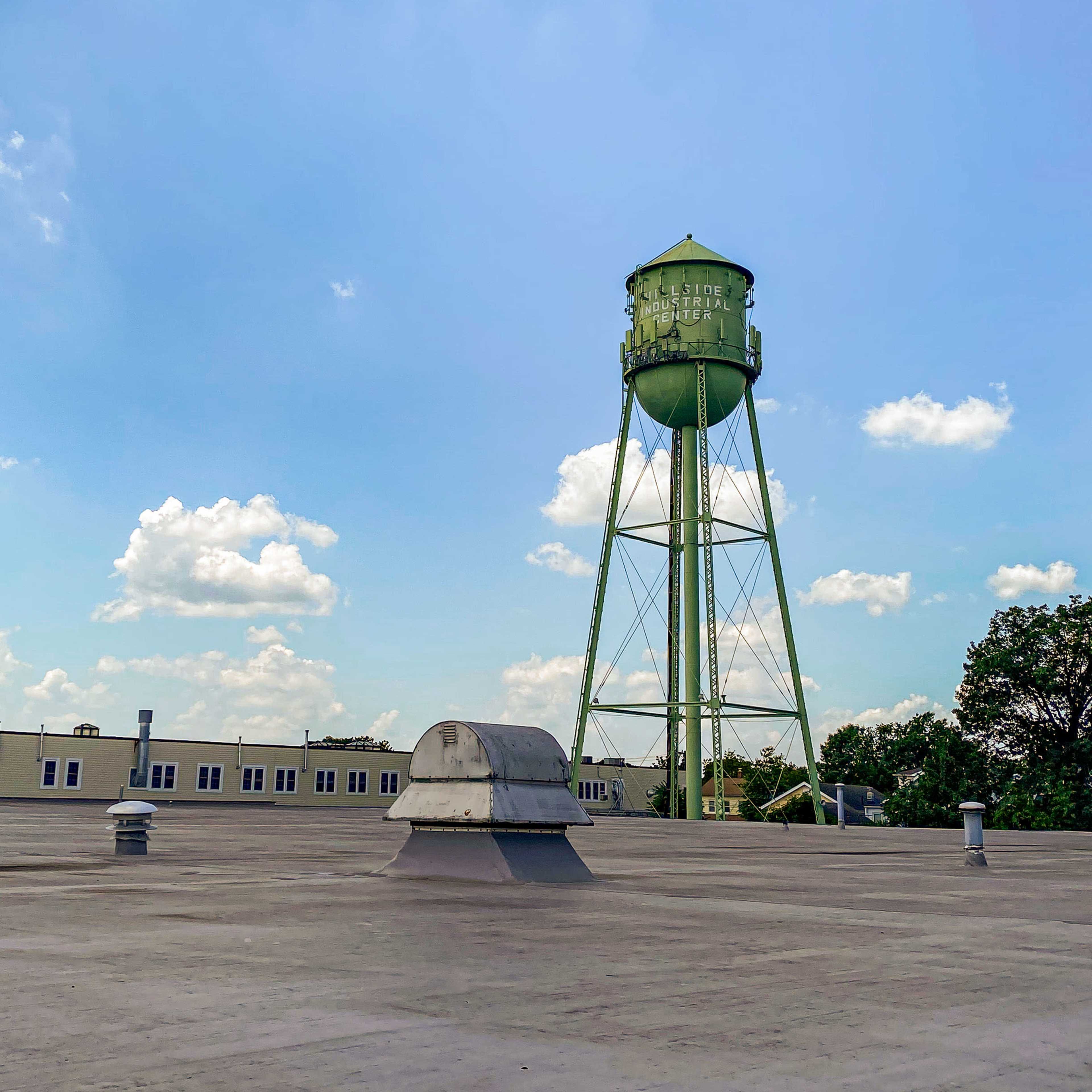 A green water tower stands tall against a blue sky, with scattered clouds and a flat rooftop in the foreground.