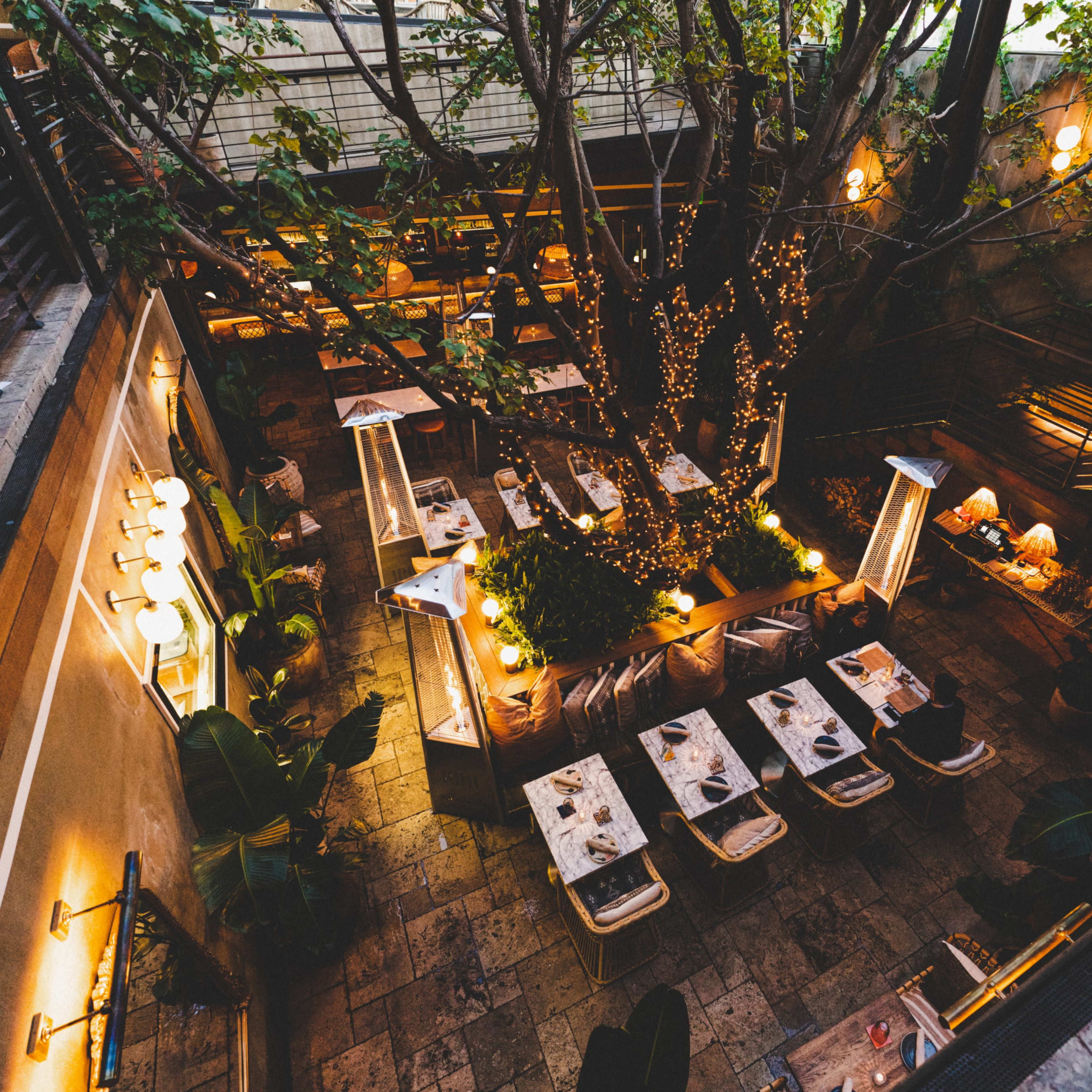 A sunken restaurant features outdoor seating surrounded by greenery and string lights illuminating the space.