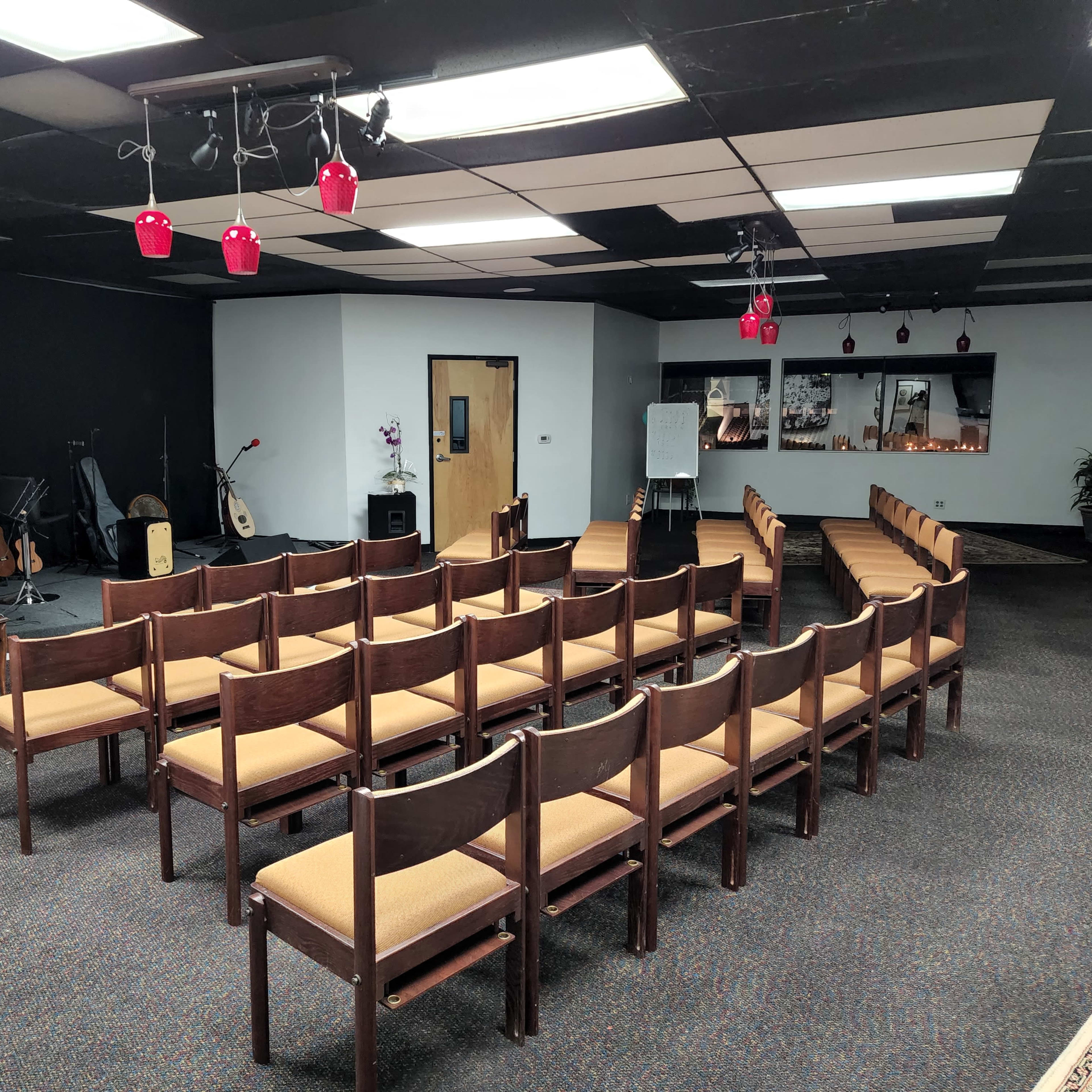 A room arranged with rows of wooden chairs facing a wall with a piano and musical instruments visible in the background.