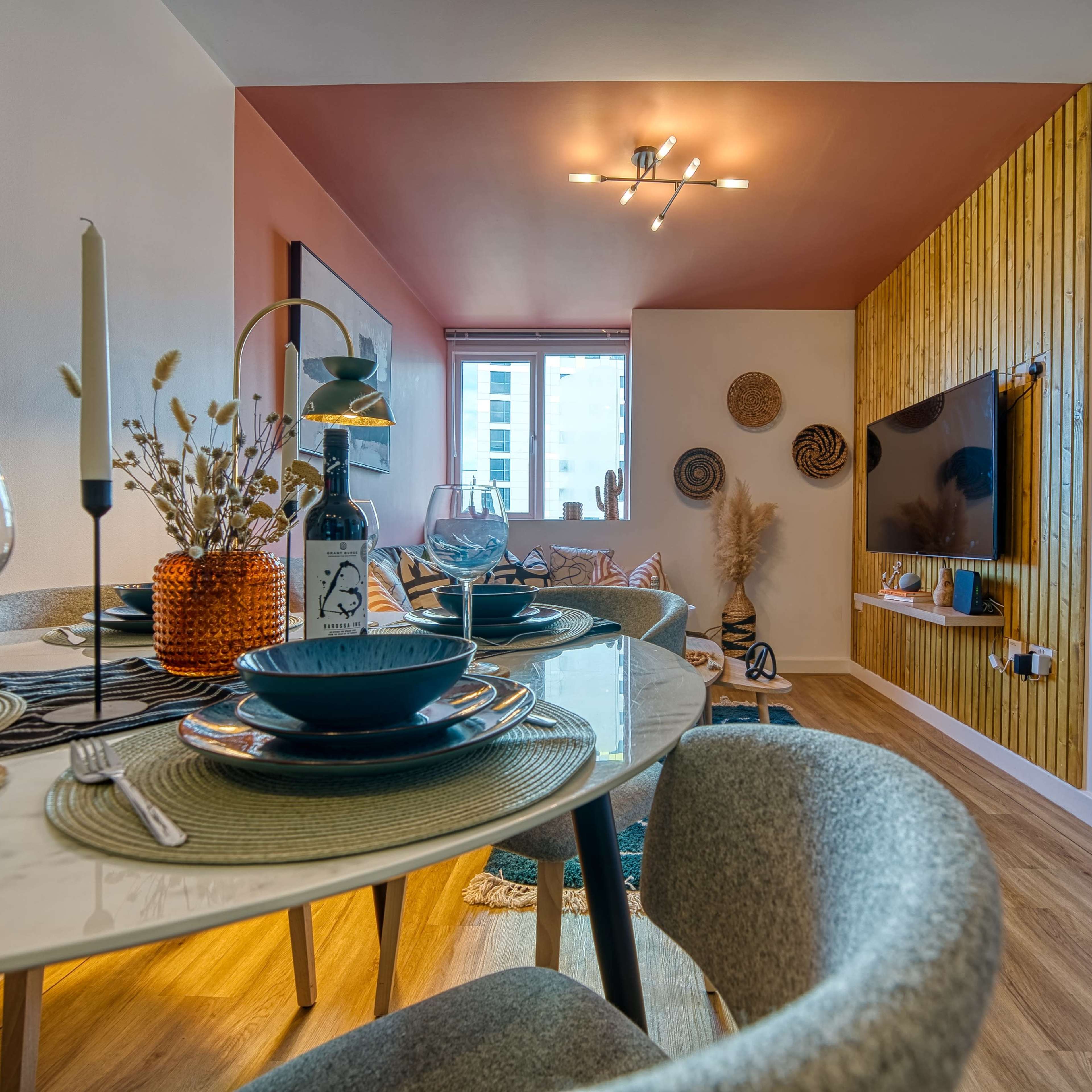A modern dining area with a round table set for a meal, leading into a cozy living space featuring a television and decorative wall elements.