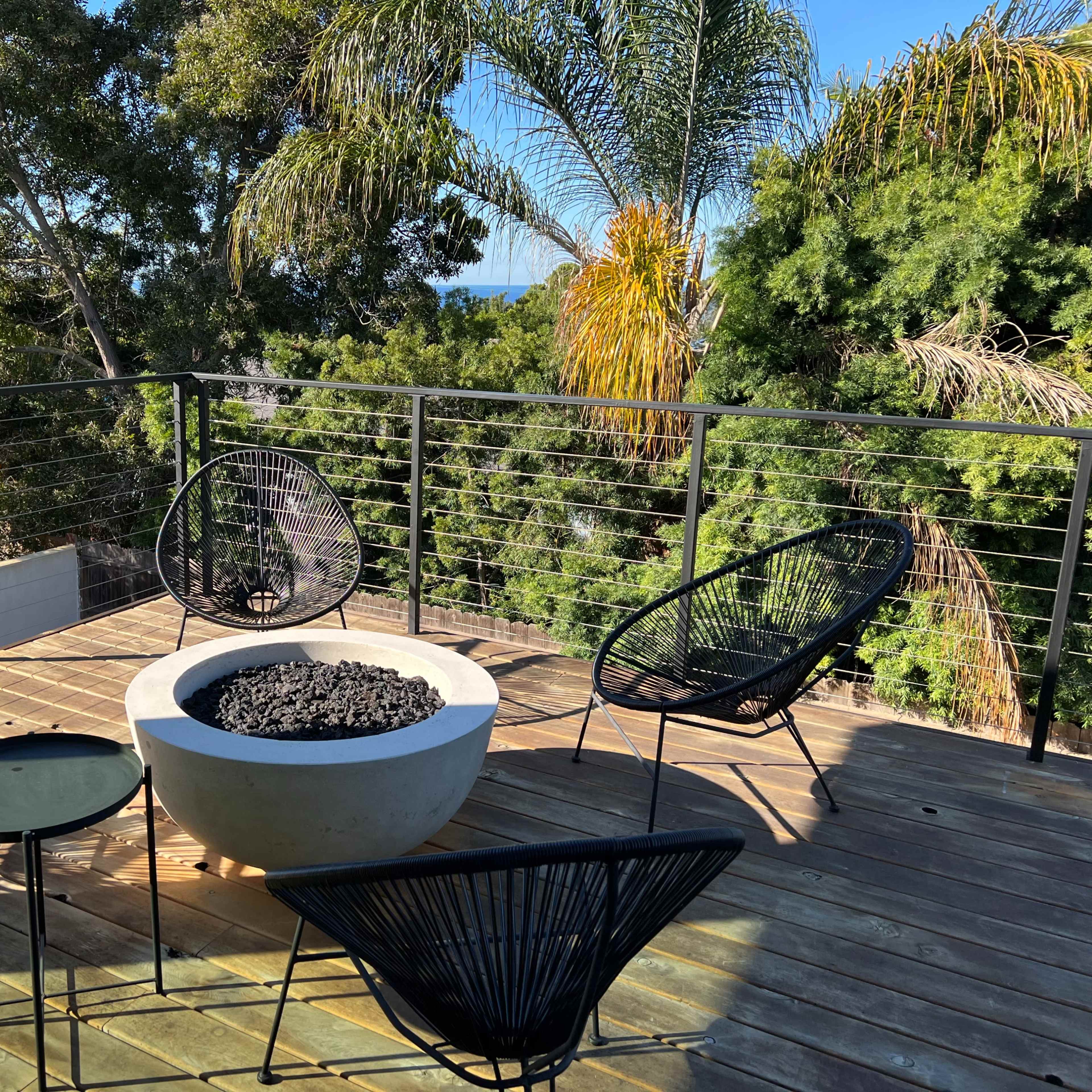 A modern outdoor seating area features four black wire chairs arranged around a circular fire pit on a wooden deck, with lush greenery and palm trees in the background.