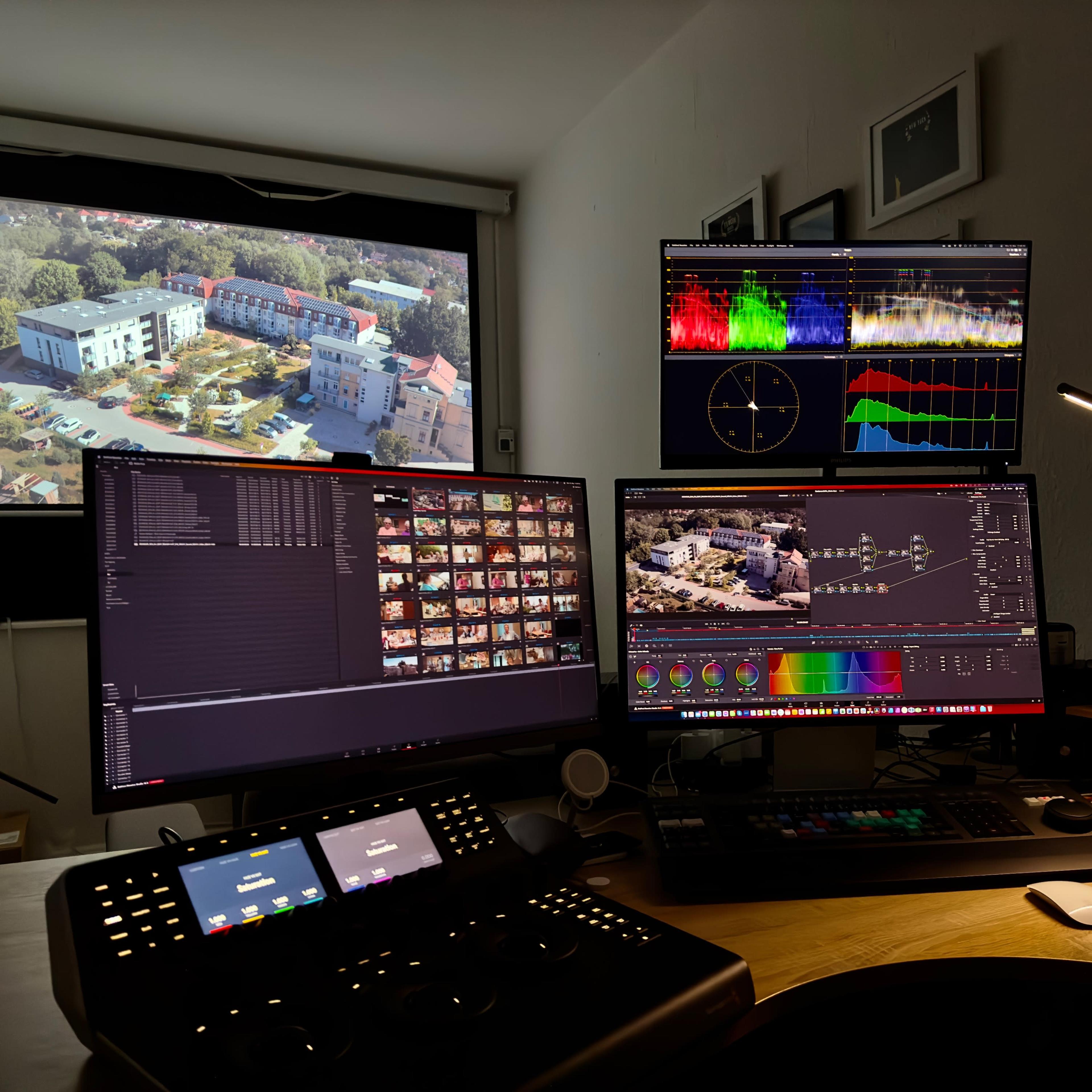 A digital editing setup features multiple monitors displaying video footage and color grading tools, with a view of a building outside the window.