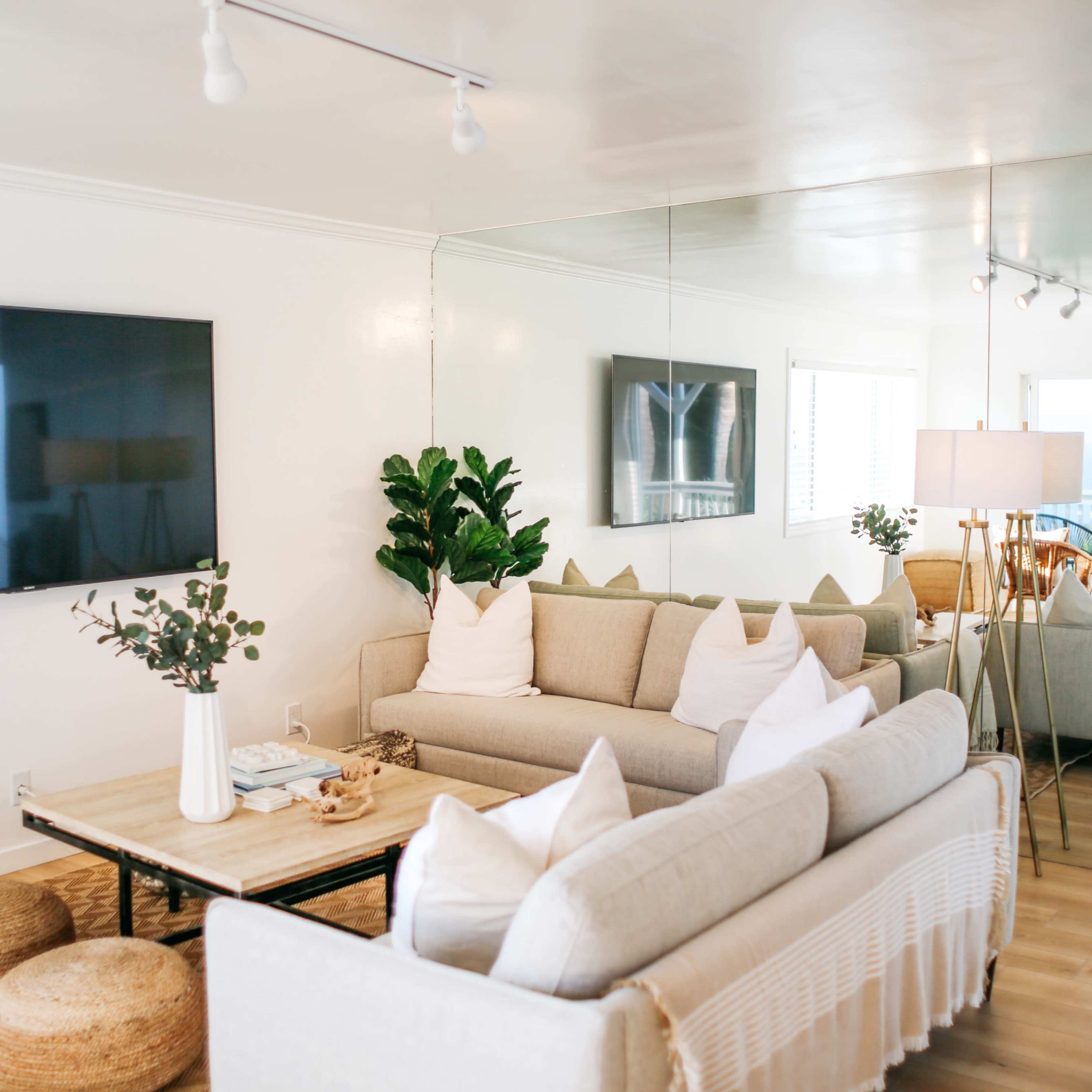 A modern living room with a light color scheme, featuring two sofas, a coffee table, and a large wall-mounted TV, along with plants and decorative elements.