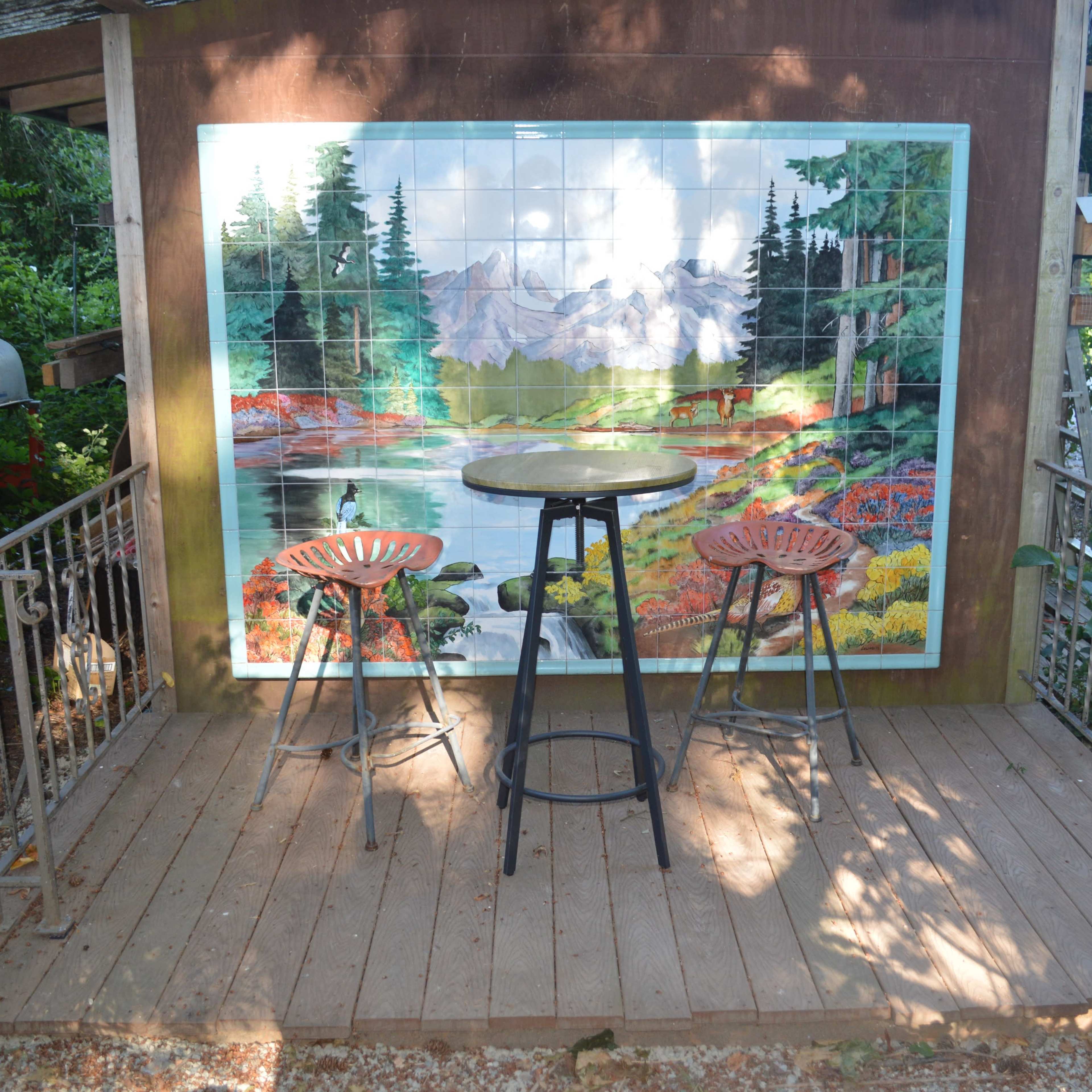 A wooden deck with two stools faces a mural of a natural landscape, surrounded by greenery.