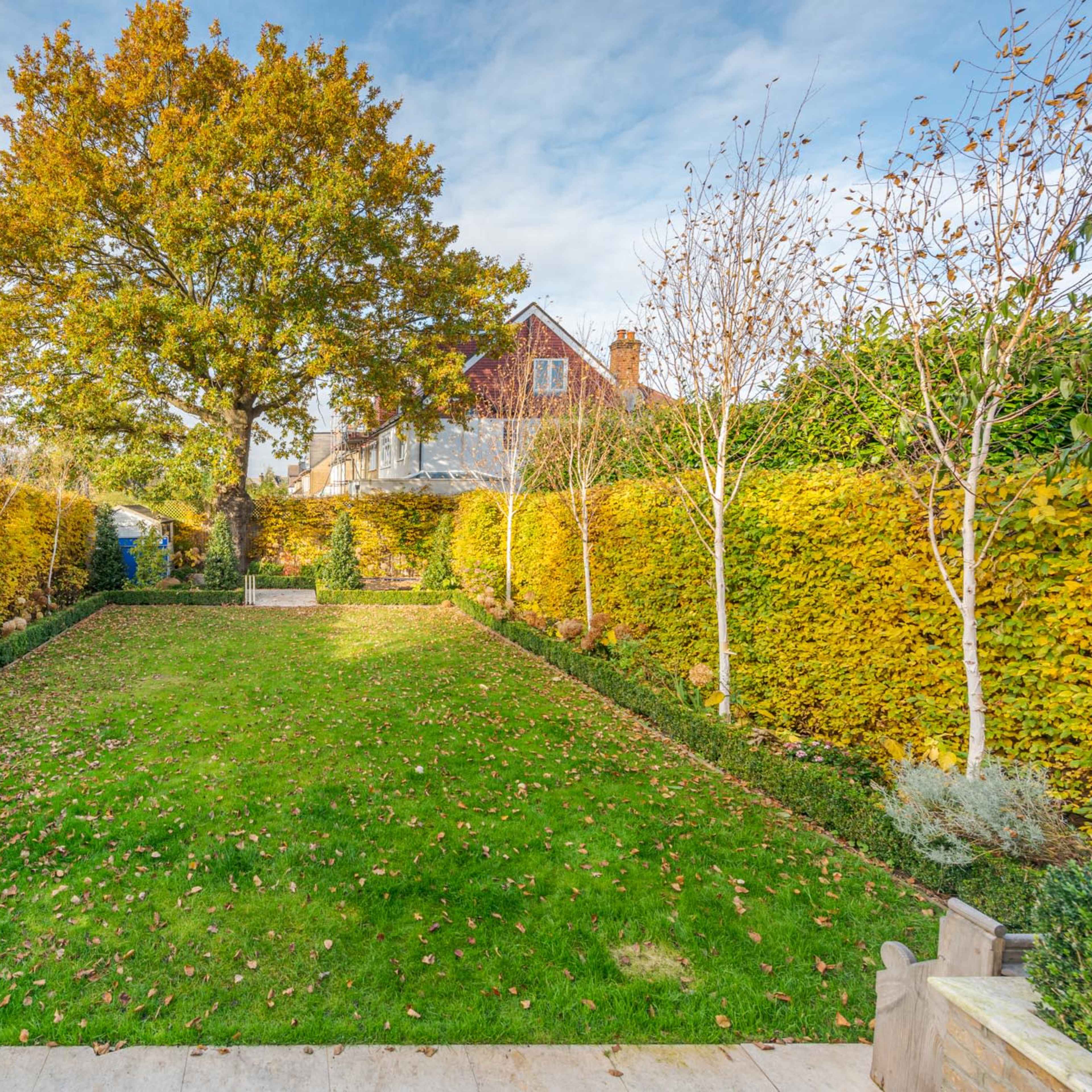 A symmetrical garden features a large tree at the center, surrounded by neatly trimmed hedges and pathways, with autumn foliage visible on the trees.