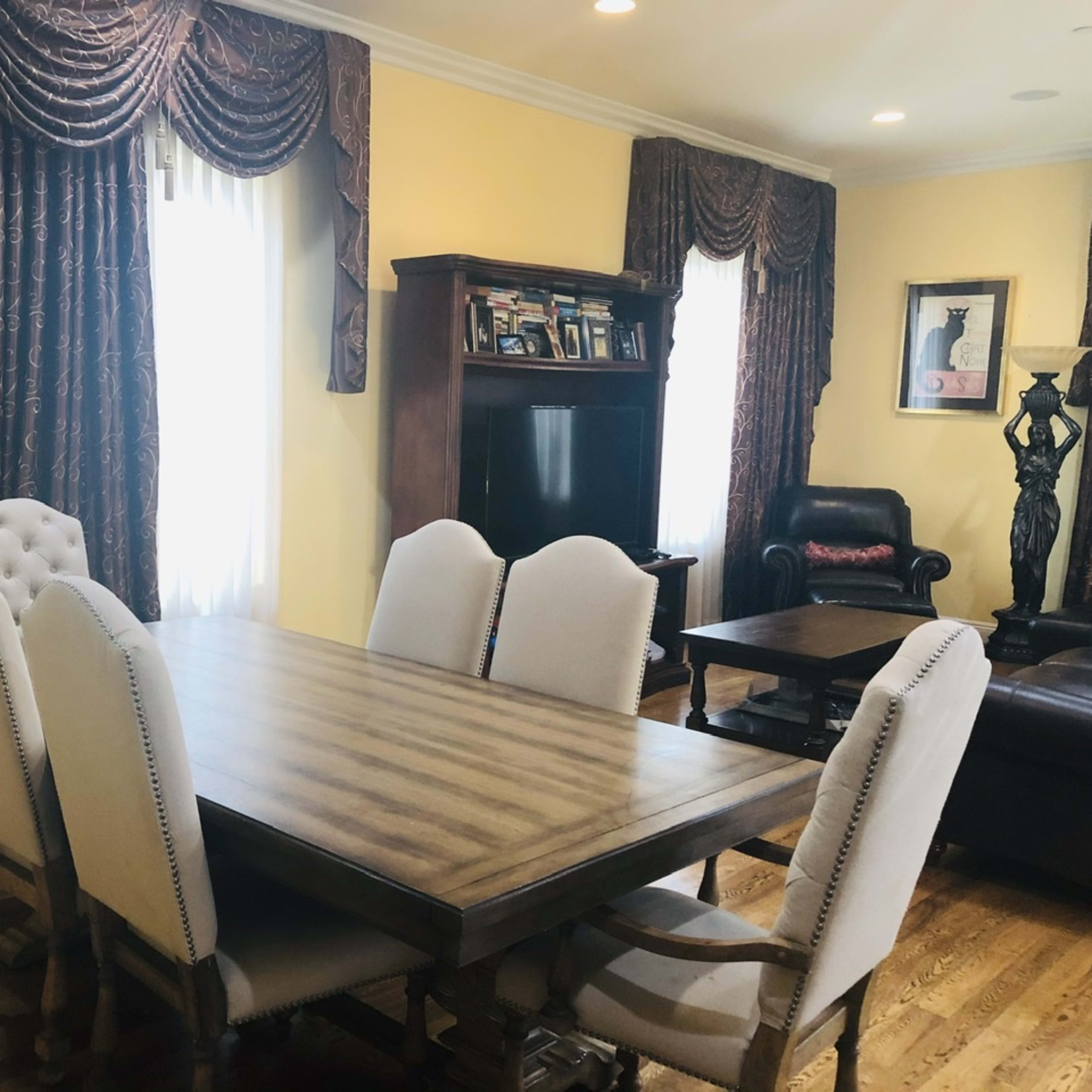 A wooden dining table with six upholstered chairs is positioned in a brightly lit room featuring yellow walls and decorative curtains.