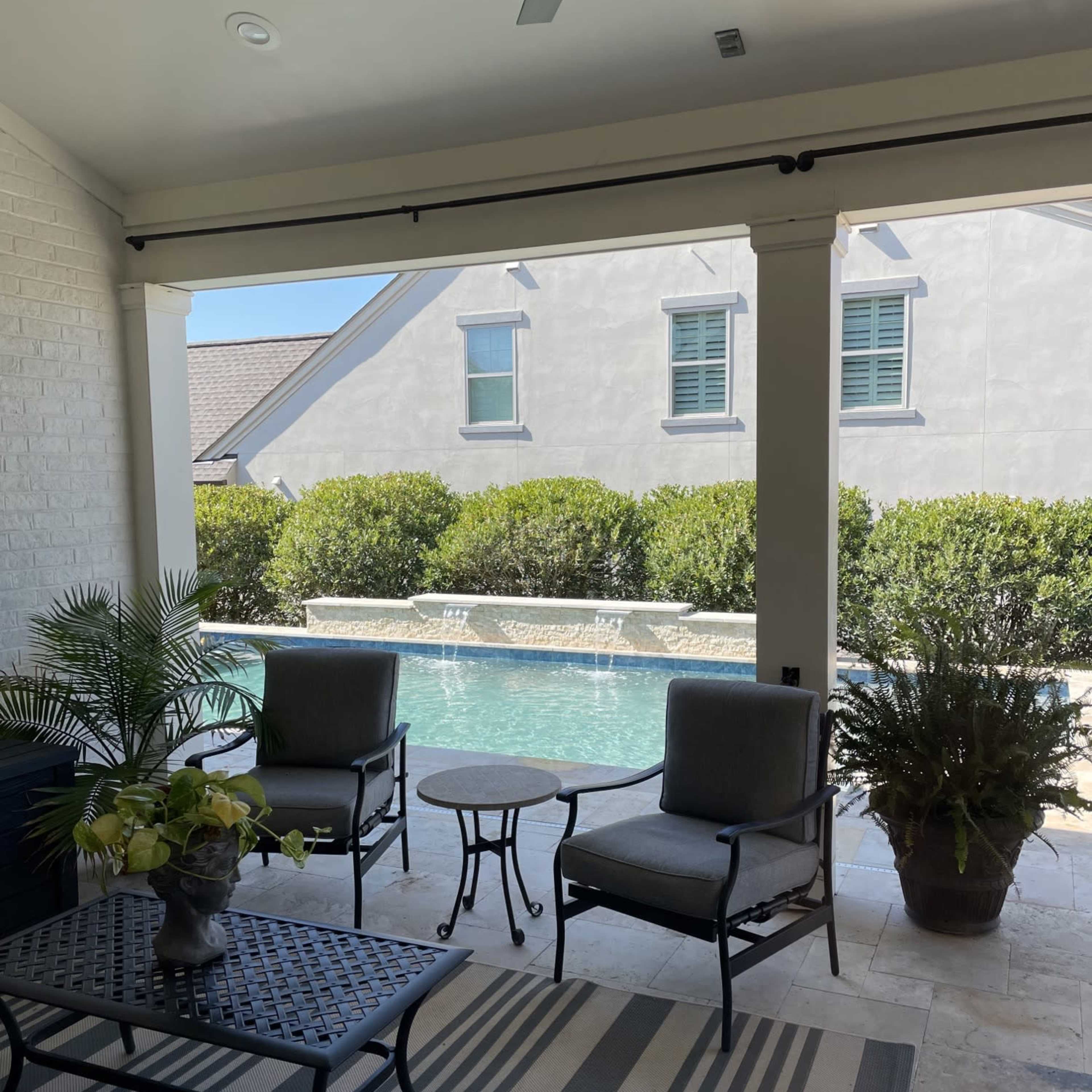 A shaded patio area features two chairs and a table, overlooking a swimming pool surrounded by greenery.