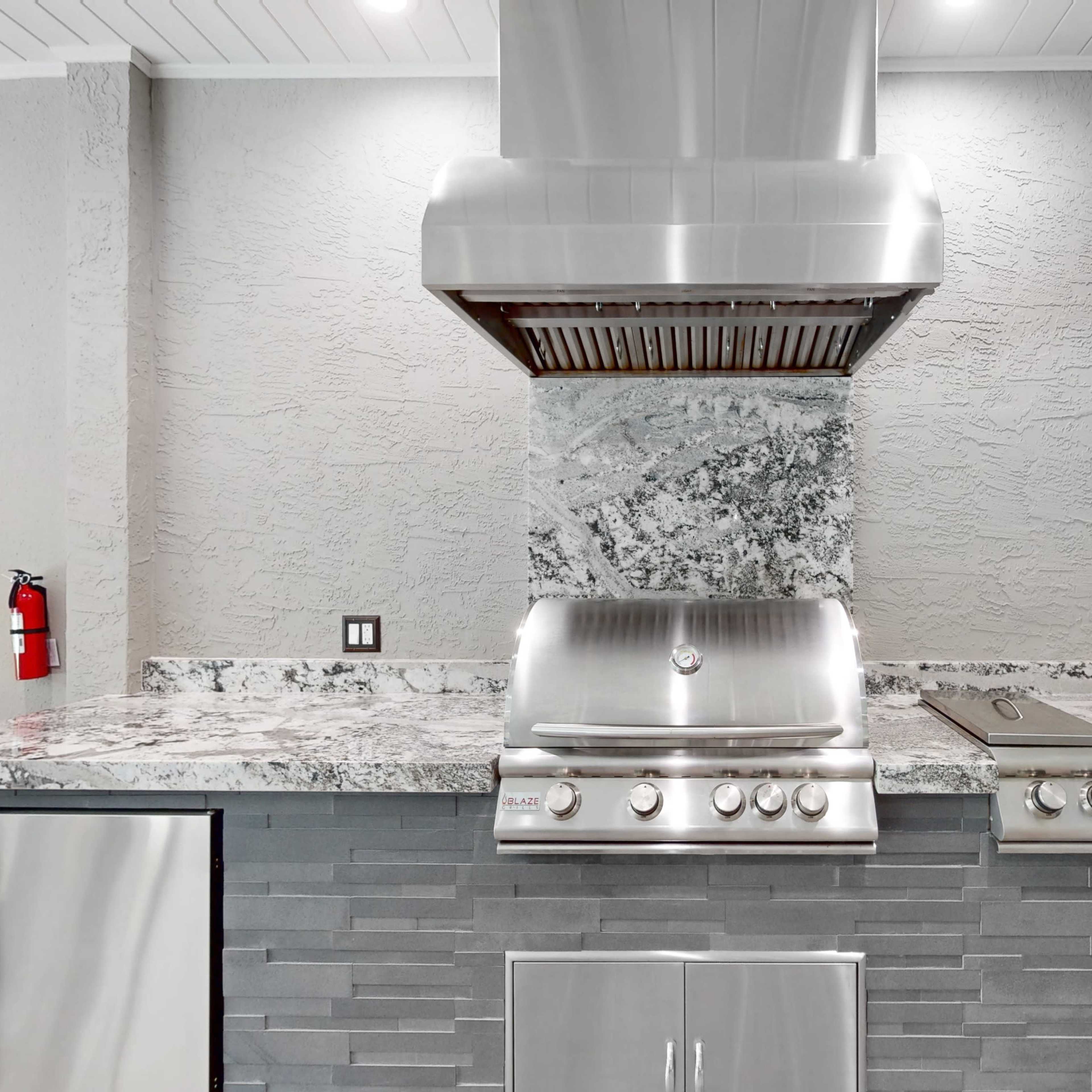 A modern outdoor kitchen with a stainless steel grill and hood, surrounded by a marble countertop and gray stone walls.