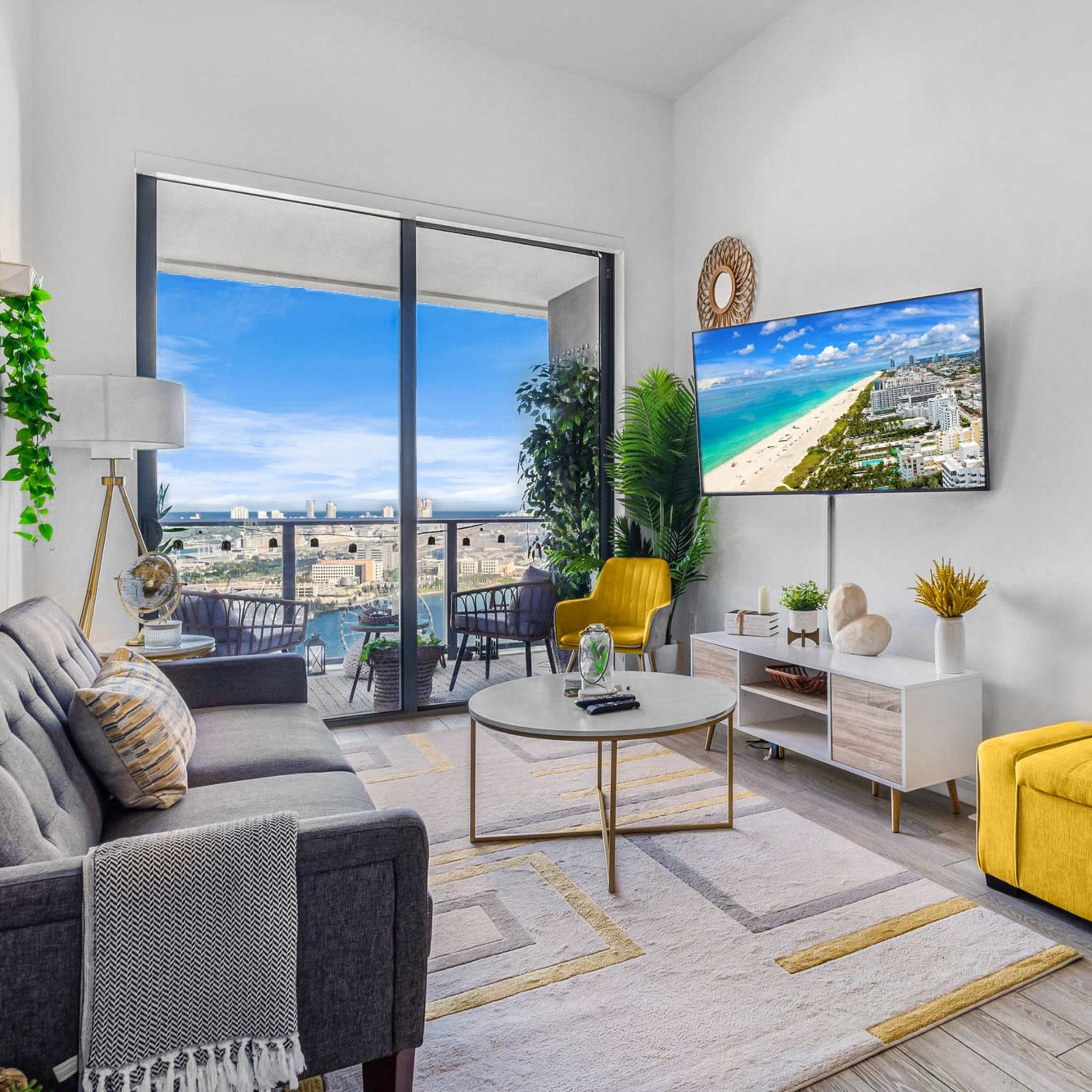 A modern living room features a gray sofa, yellow accent furniture, and a large TV displaying a beach view.