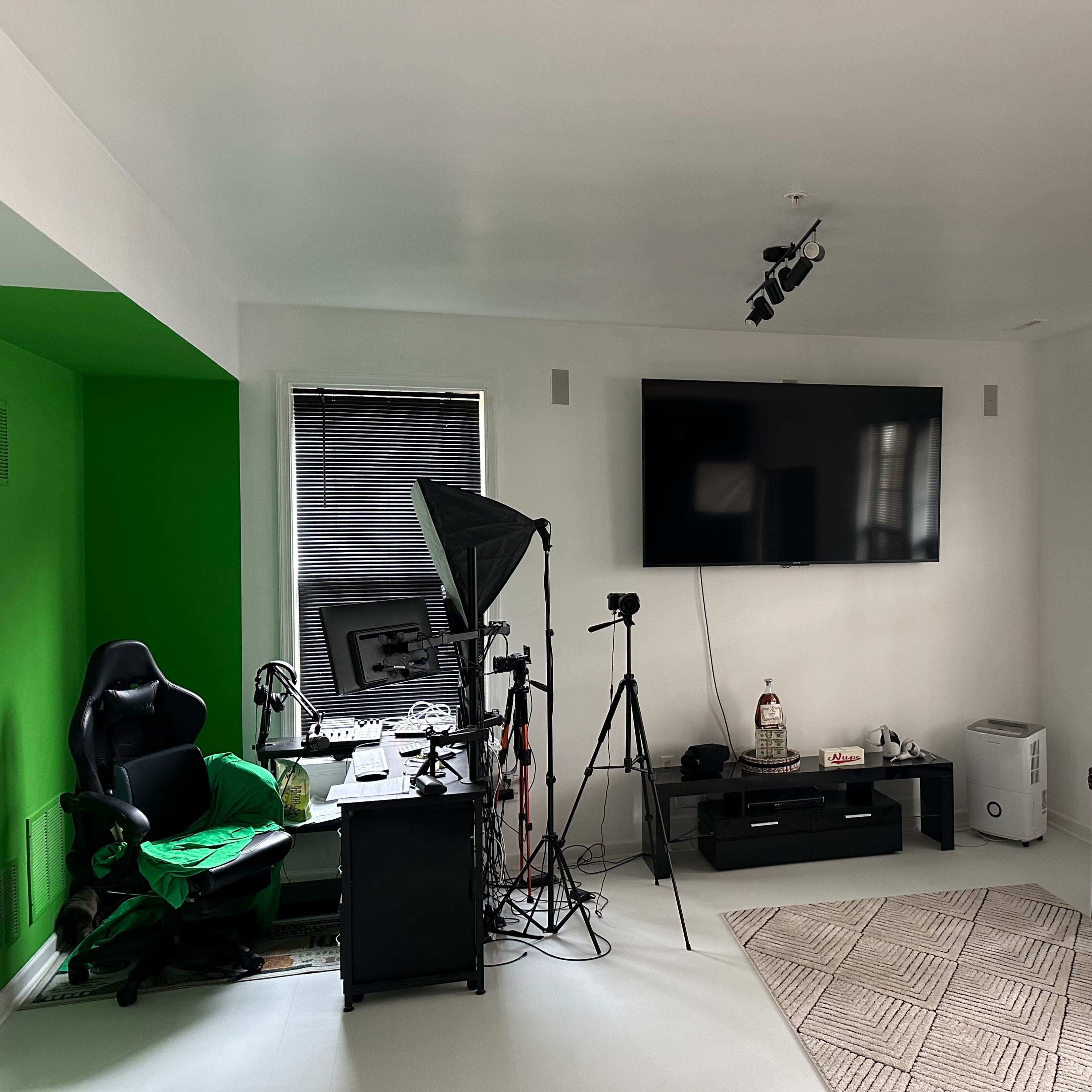 A room features a green accent wall, a black desk with a computer setup, a camera on a tripod, and two chairs near a television mounted on the wall.