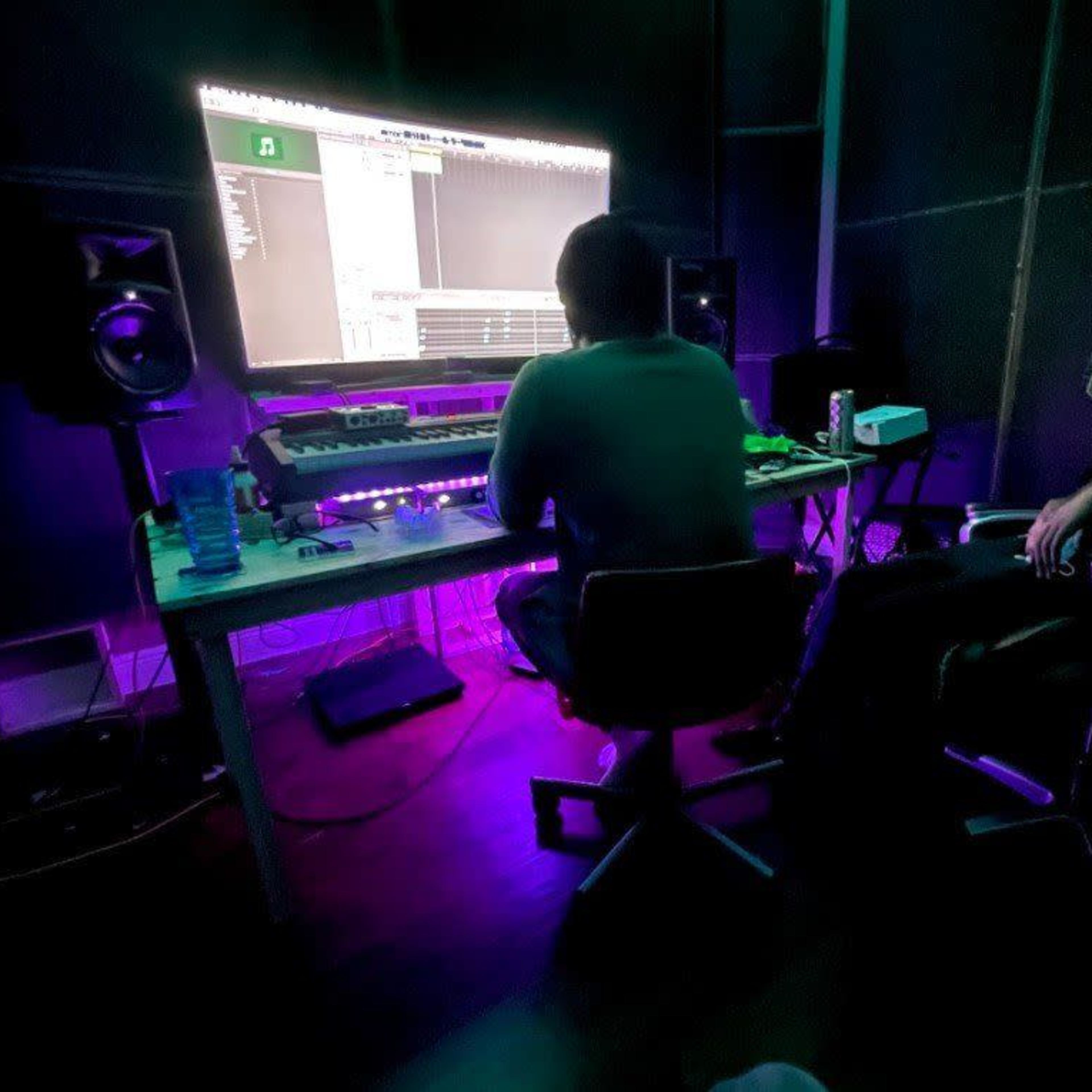 Two individuals are seated in a dimly lit room with a large computer monitor displaying music software, surrounded by audio equipment.