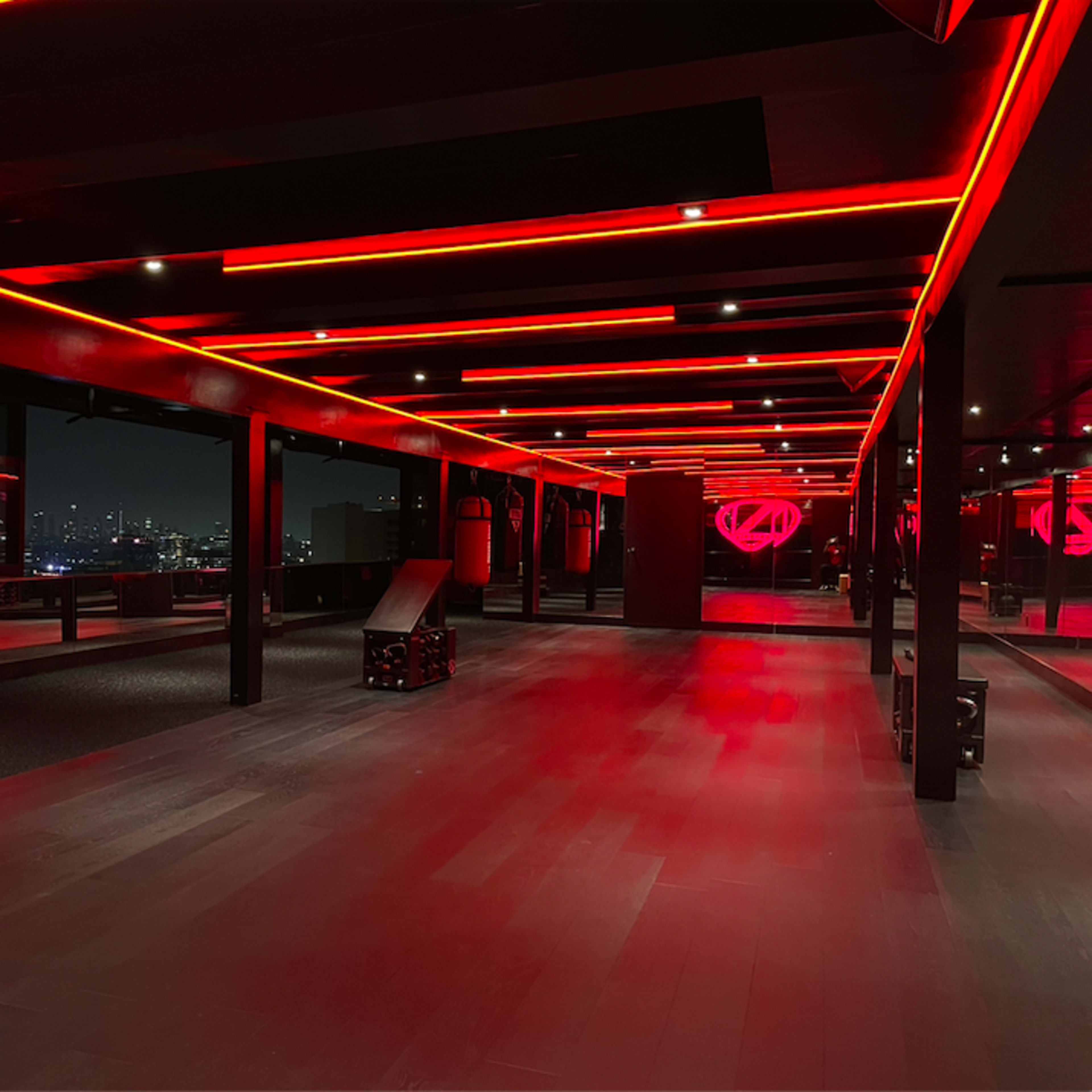 The interior of a modern building features wide wooden floors and walls illuminated by red neon lights, with large windows revealing a city skyline at night.