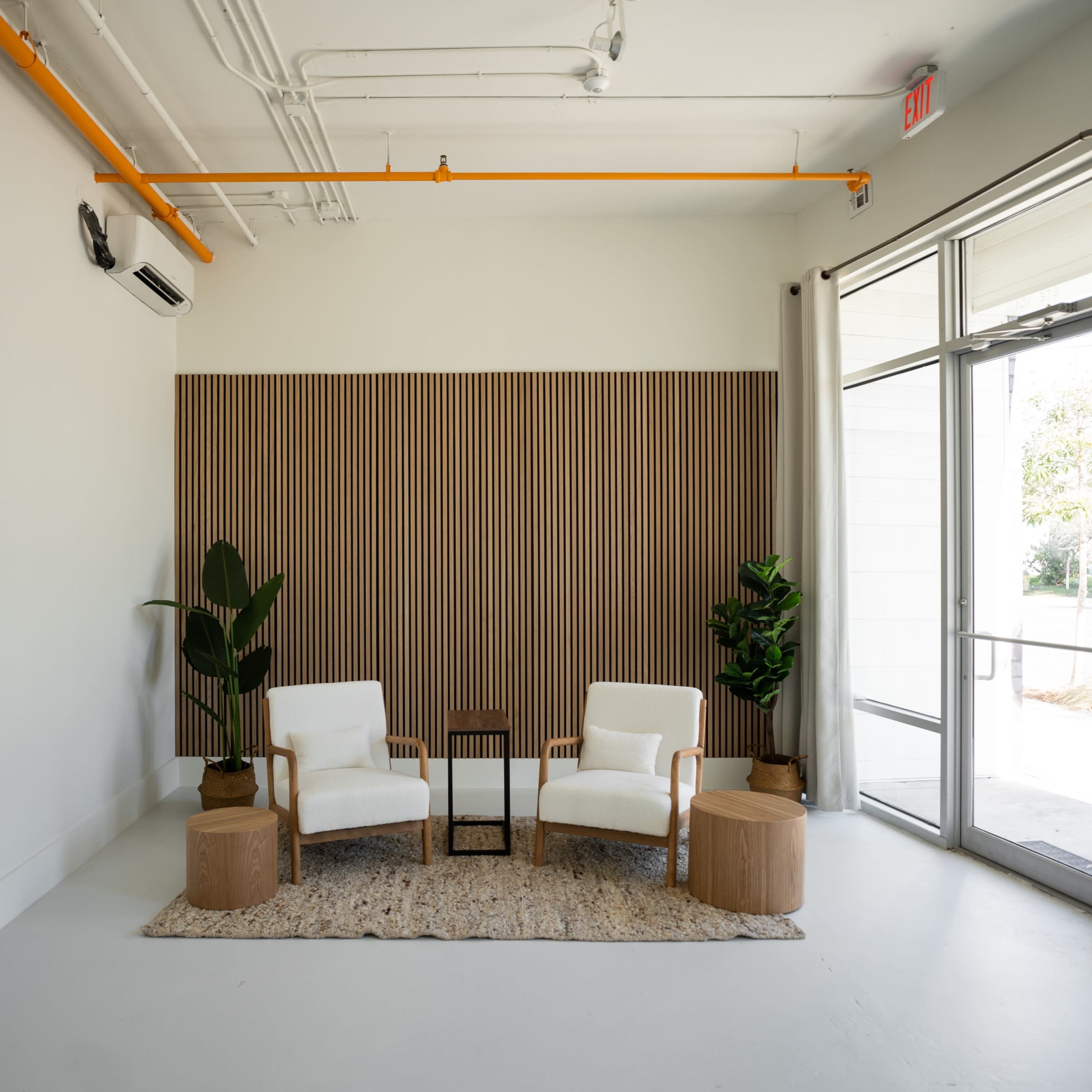 A minimalist interior space with two white chairs, a small table, and plants beside a wood panel backdrop.