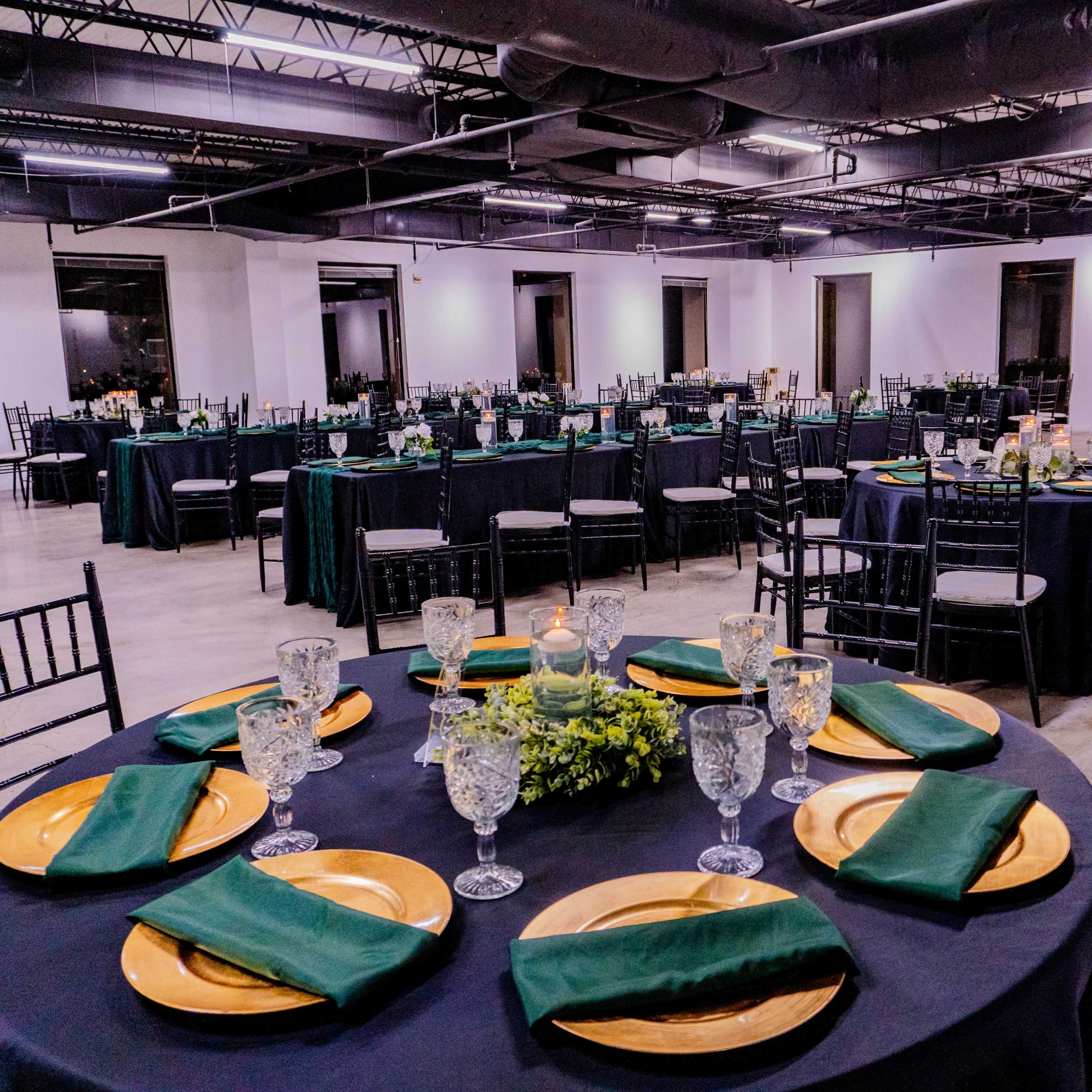 A spacious event room features round tables set with dark tablecloths, gold chargers, green napkins, and glassware, surrounded by additional tables and chairs.