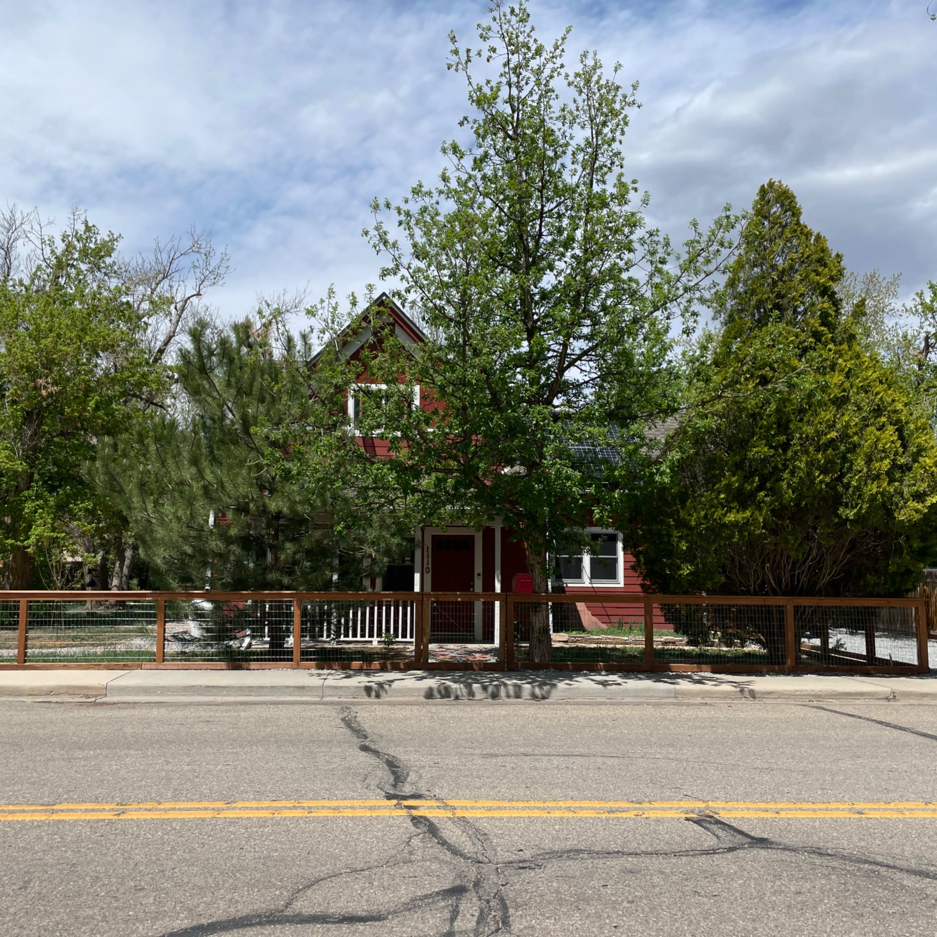 A red house with a porch is surrounded by trees and a wooden fence along a cracked road.