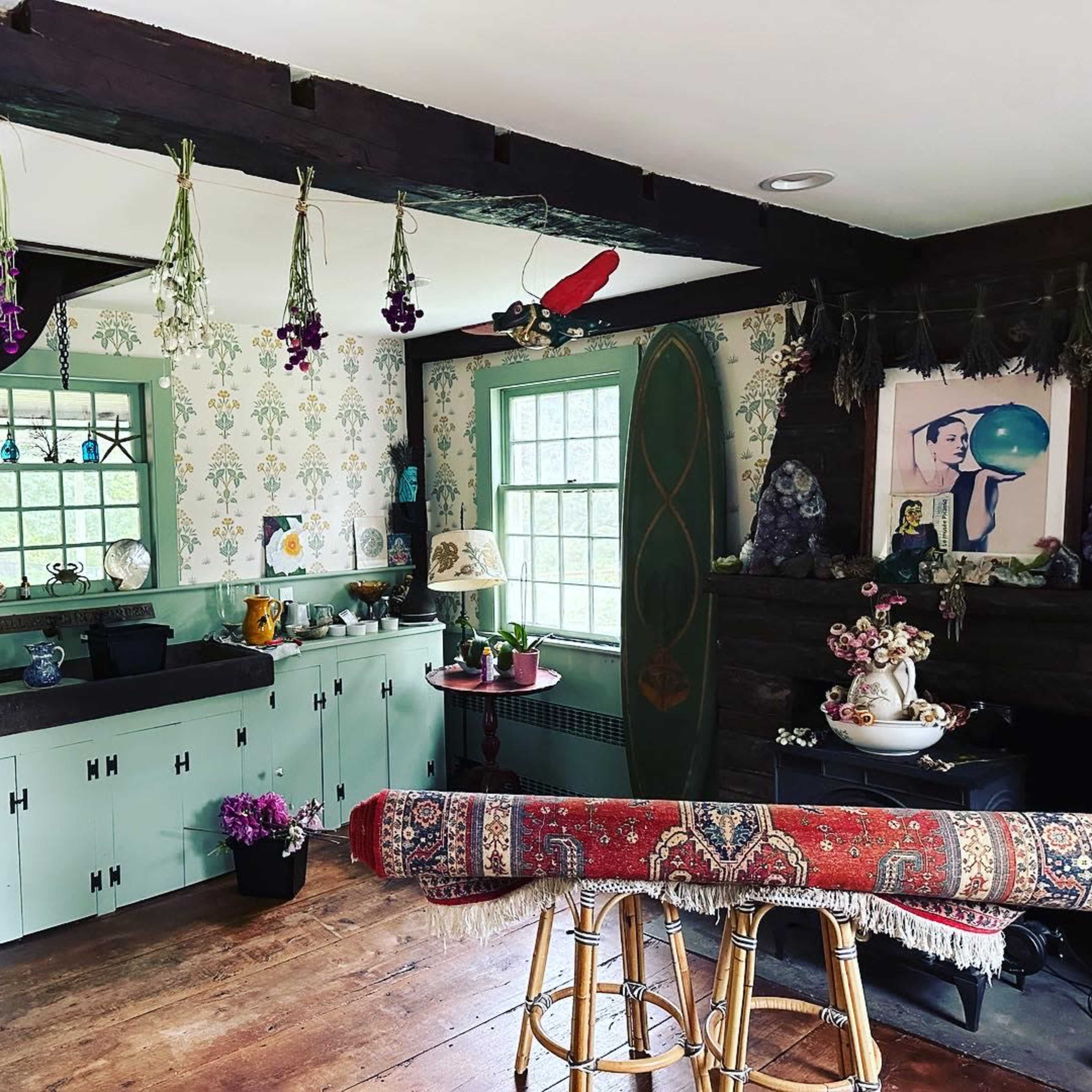 A cozy, eclectic room with green-trimmed windows, hanging dried flowers, and a mix of decorative items on shelves and furniture.