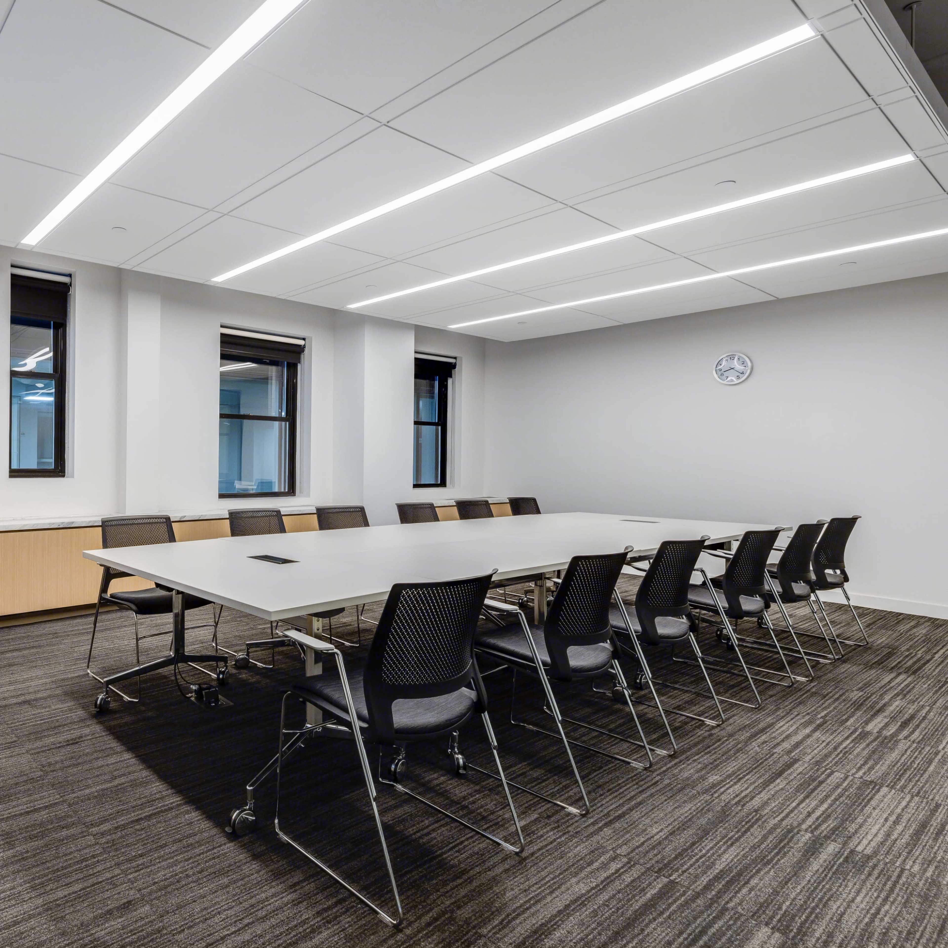 A conference room features a long table surrounded by chairs, with large windows and overhead lighting.