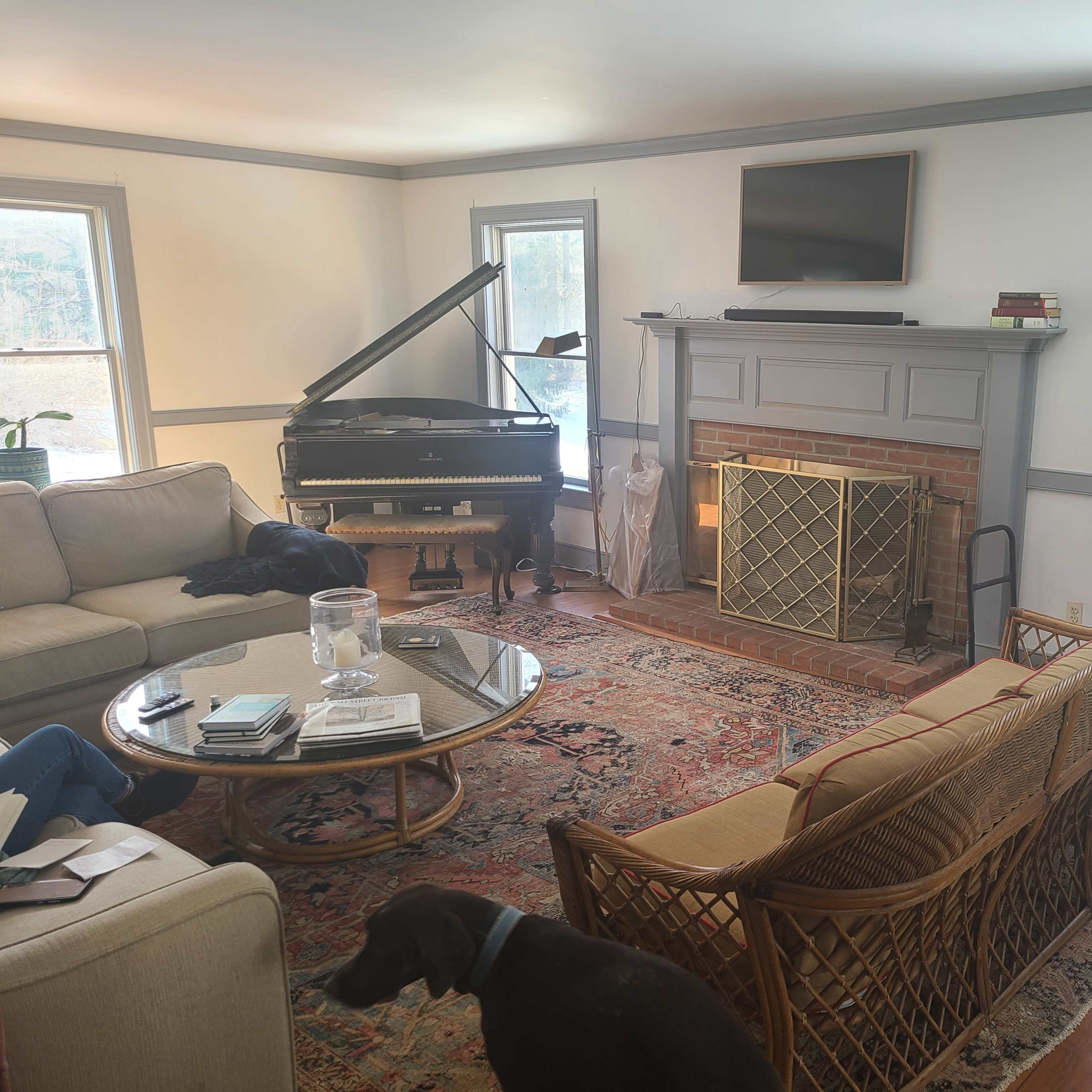 A cozy living room with a grand piano, a seating area featuring a light-colored sofa and a round coffee table, a fireplace, and large windows letting in natural light.