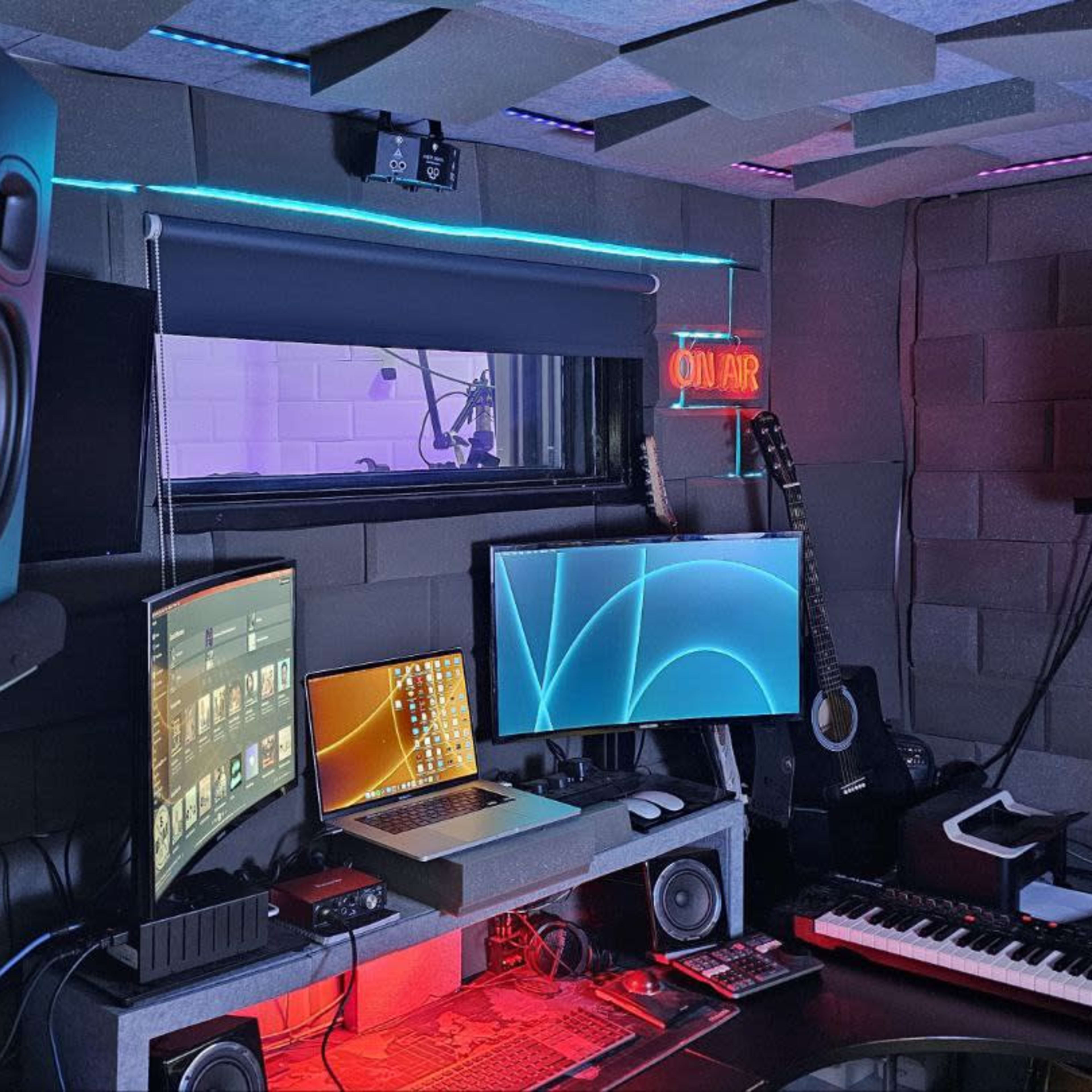 A home music production studio features multiple monitors, a keyboard, studio speakers, and acoustic panels on the walls.