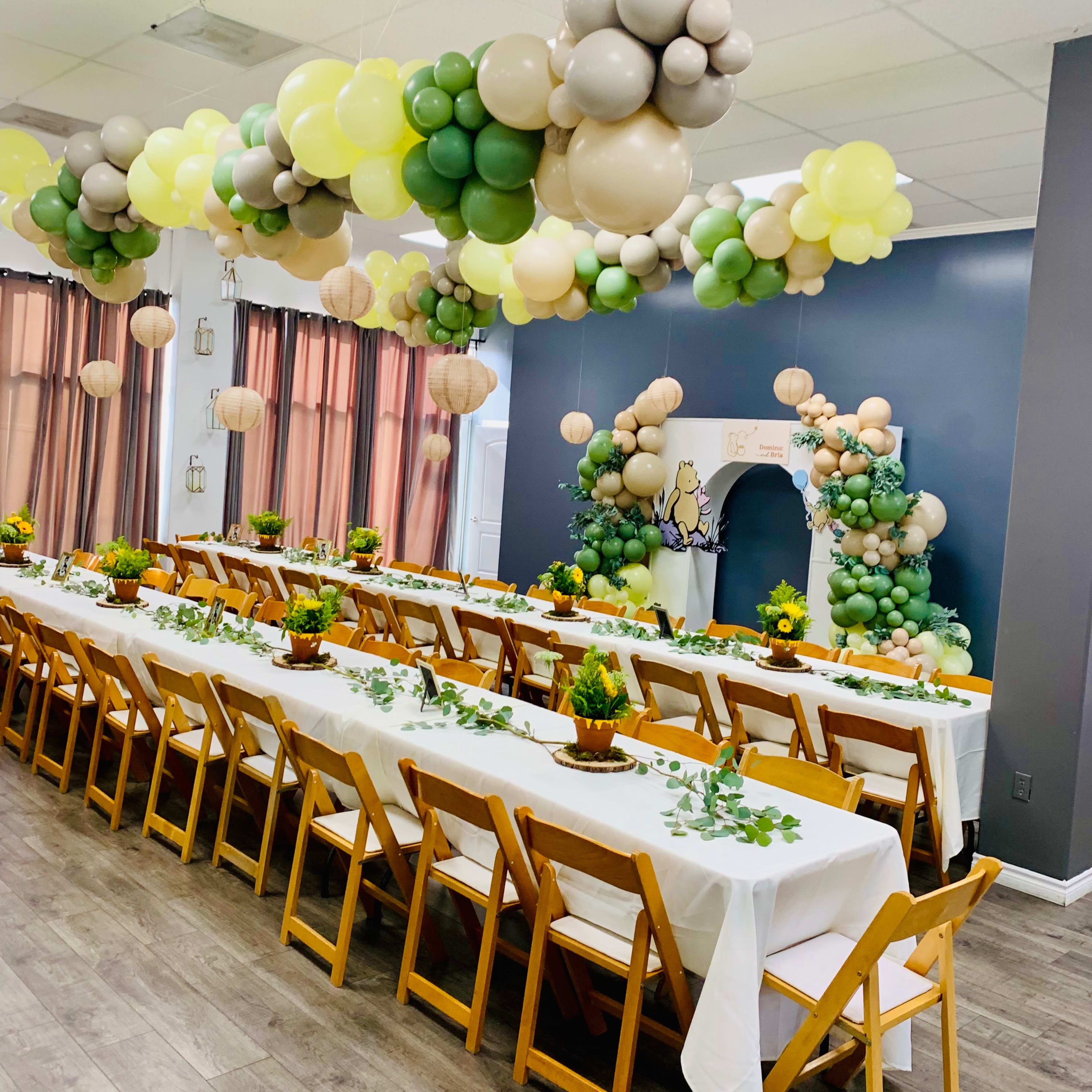 A spacious event room features long tables set with white tablecloths, surrounded by wooden chairs, and adorned with balloon decorations in various shades hanging from the ceiling.