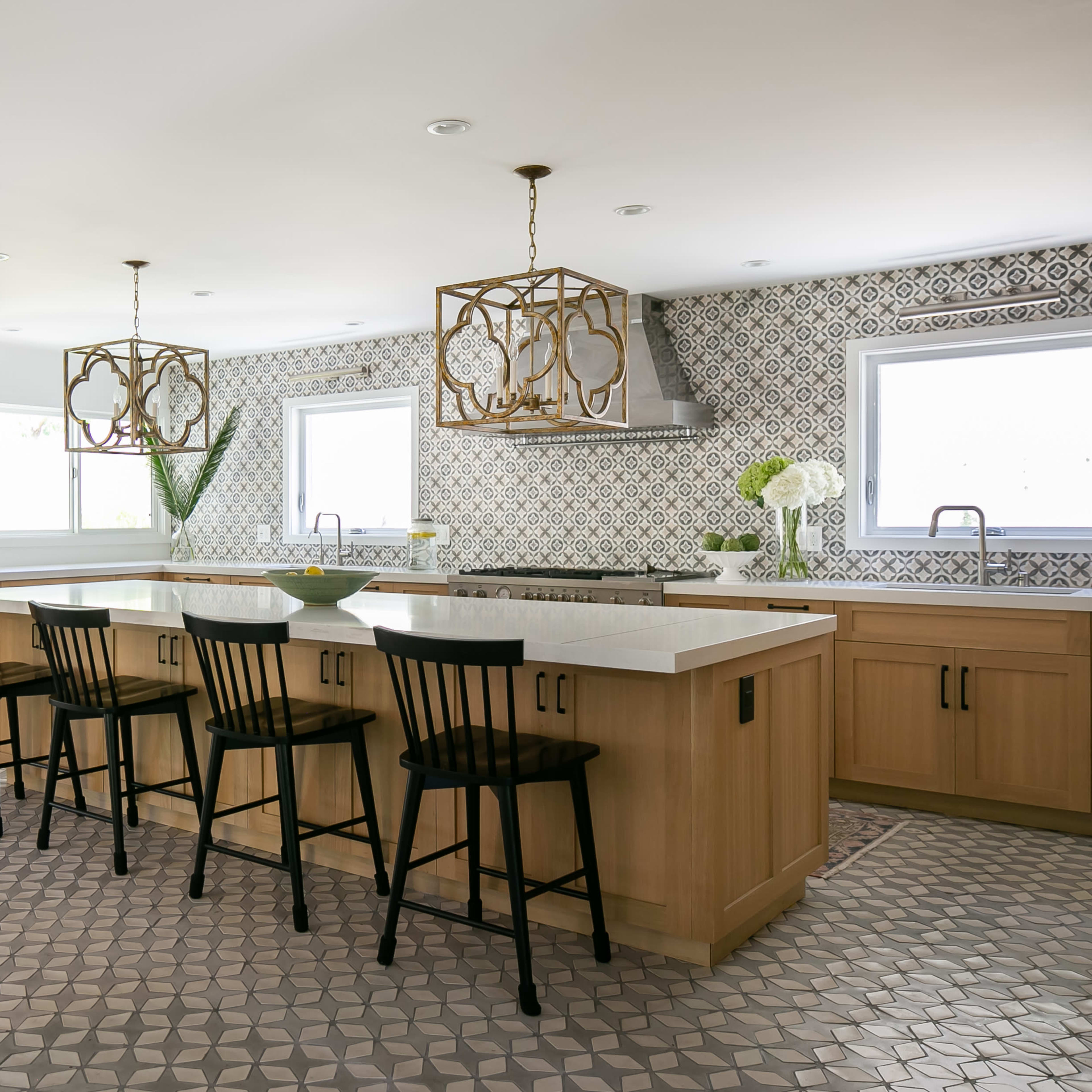 A spacious kitchen features an island with black chairs, light wooden cabinetry, and patterned tile flooring.