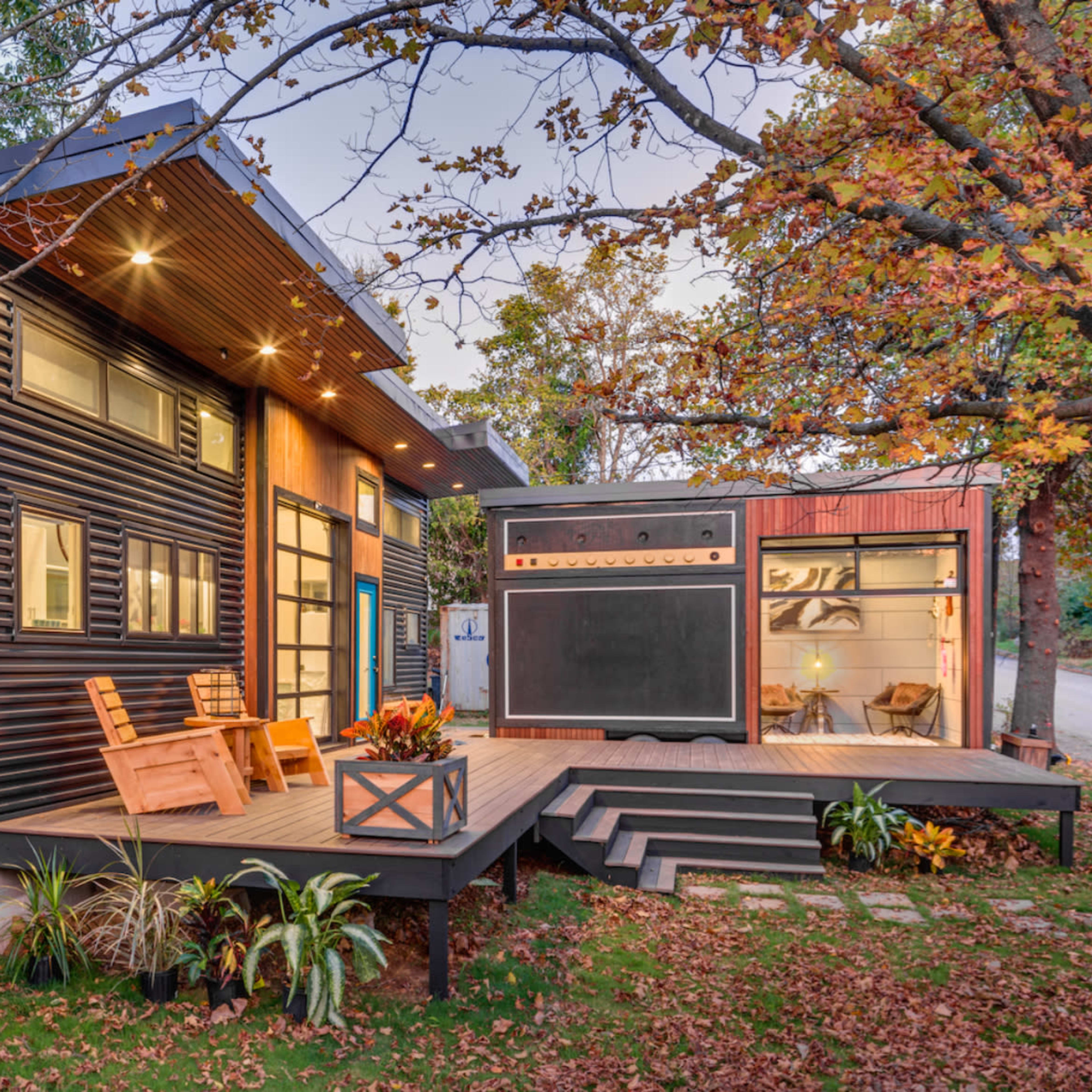 A modern house with a wooden deck and two outdoor chairs, surrounded by trees in autumn foliage.