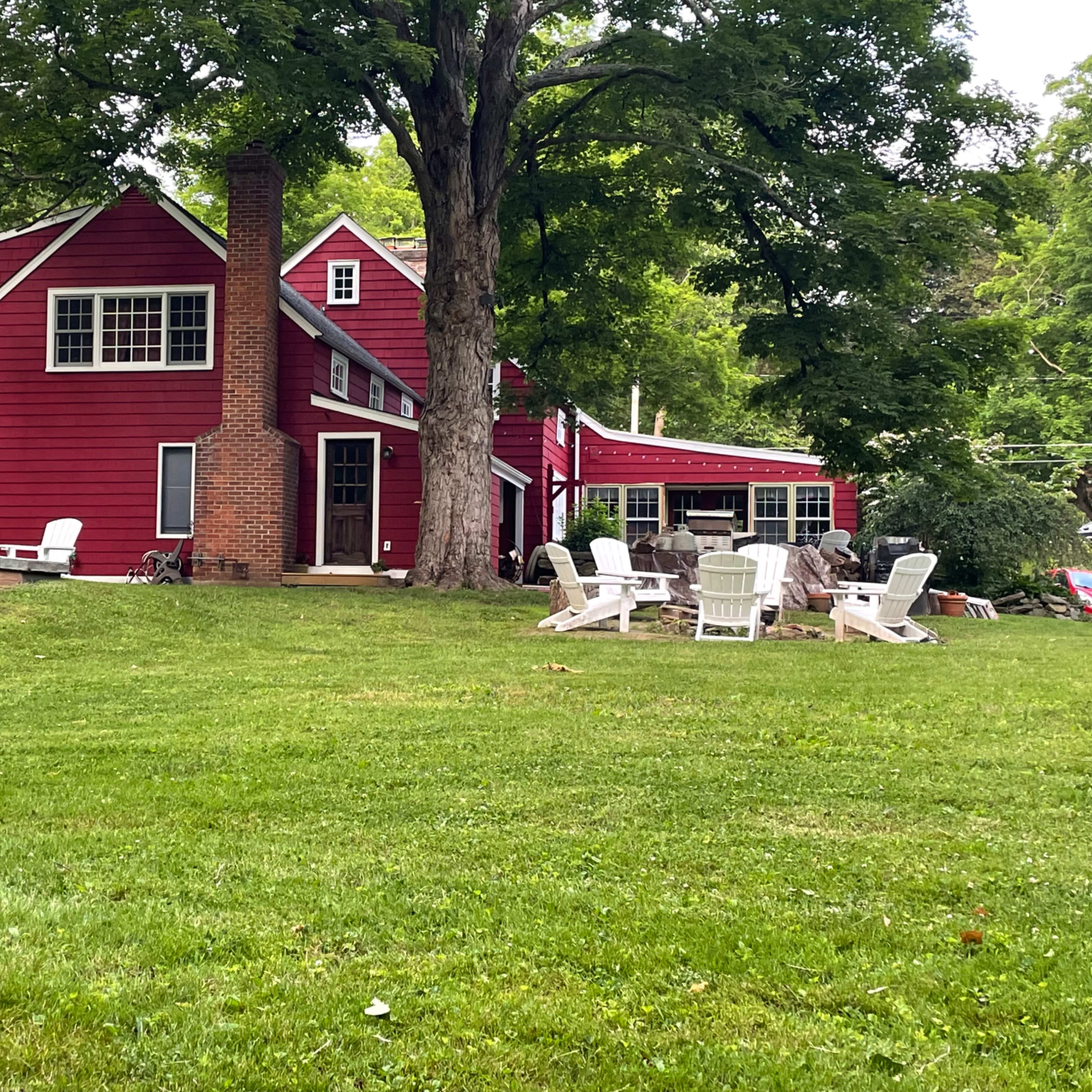 A red two-story house with a chimney and a porch, surrounded by a grassy yard and several white lawn chairs.