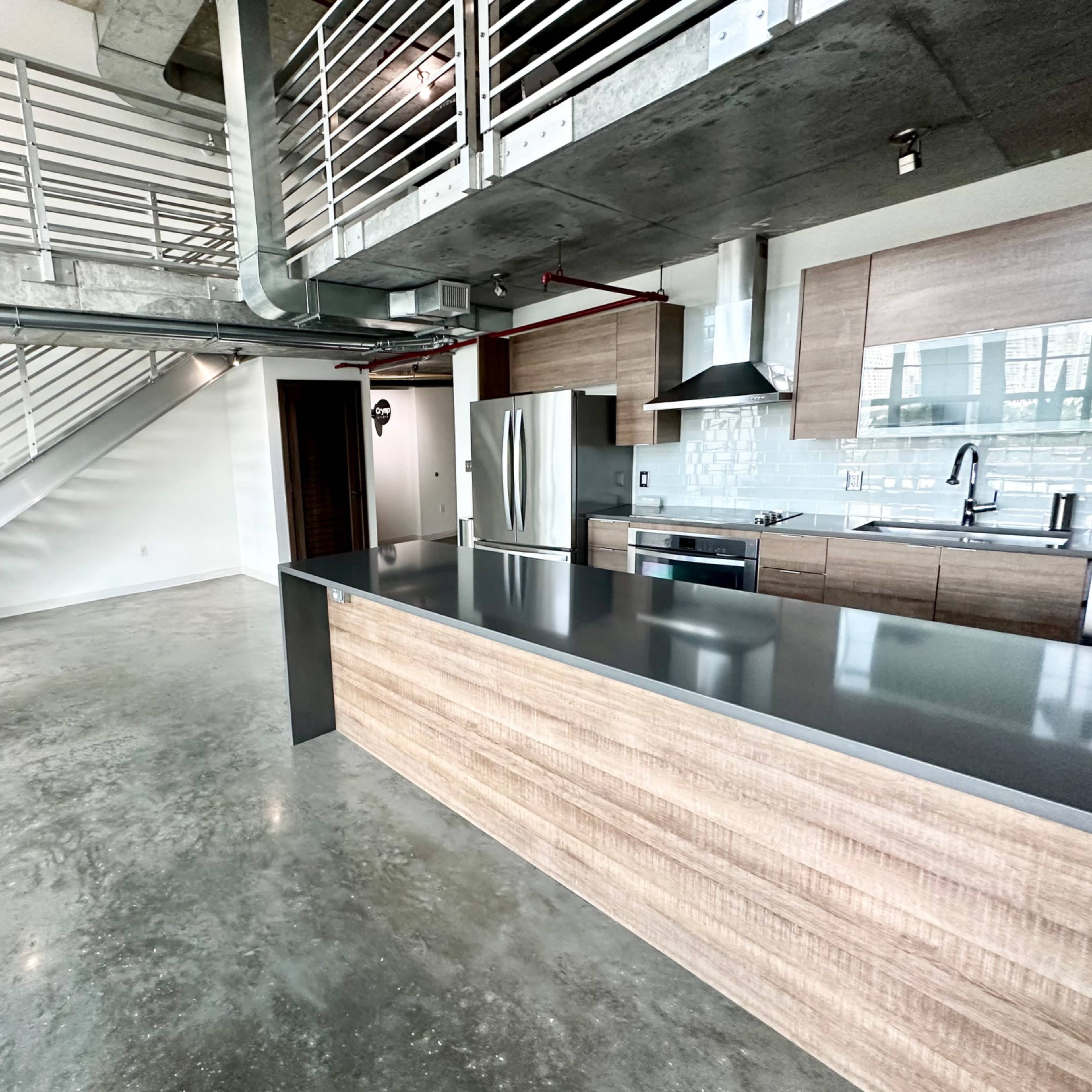 A modern, open-concept kitchen with a central island and stainless steel appliances, set in a loft-style space featuring exposed concrete floors and an industrial staircase.