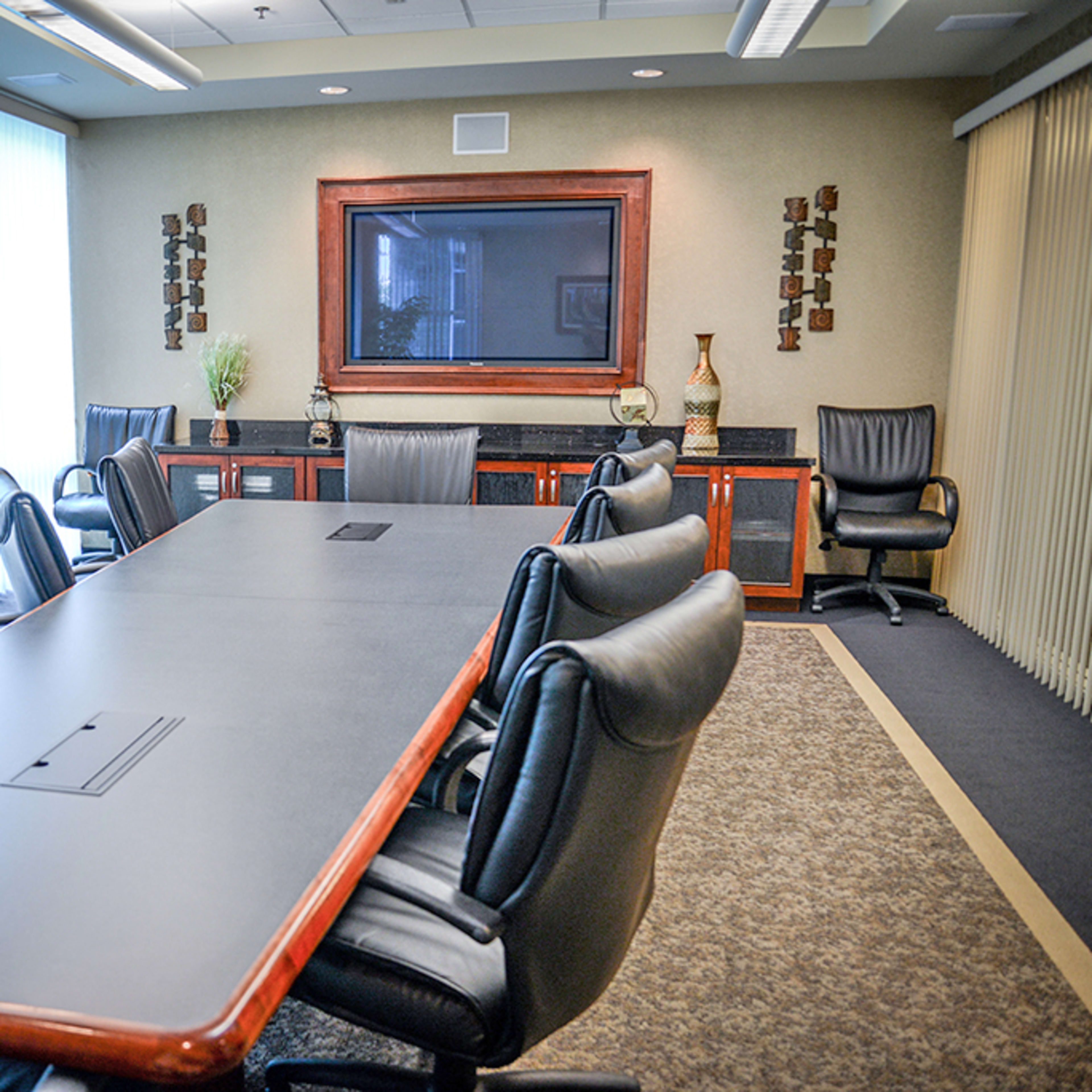 A conference room is equipped with a long table surrounded by black leather chairs and features a large screen on the wall.
