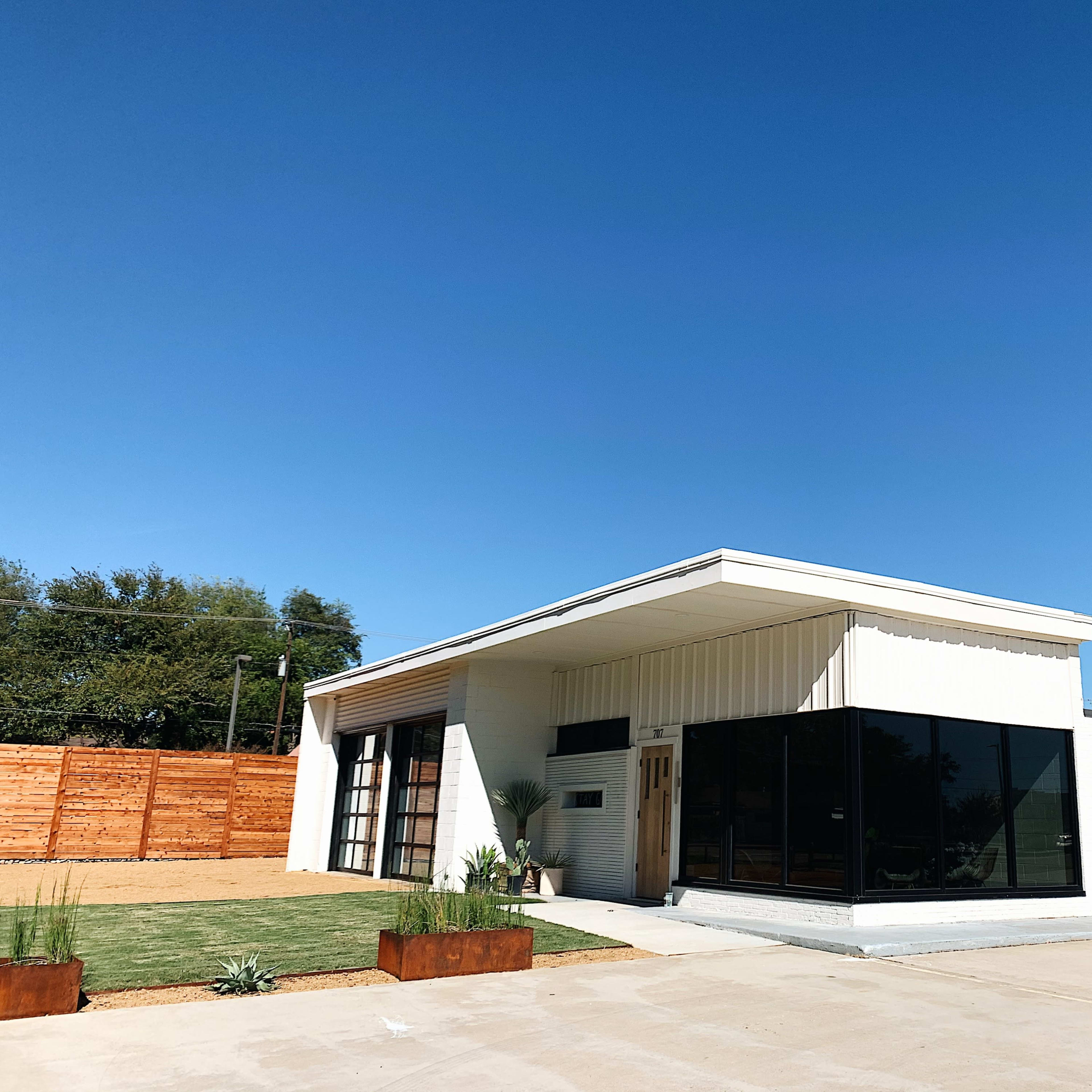 A modern single-story building with large windows and a white exterior, surrounded by a landscaped yard and a wooden fence.