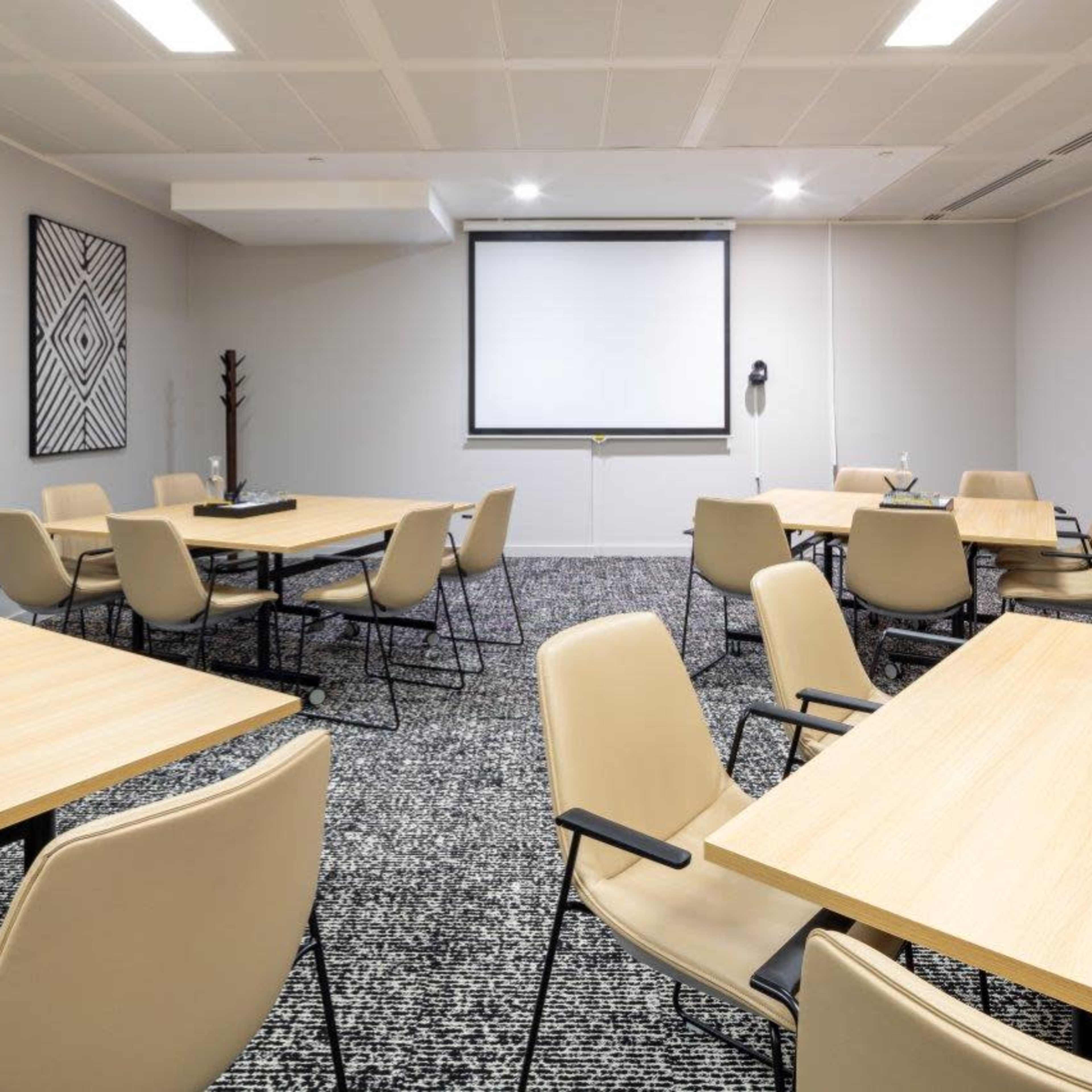 A modern meeting room with several wooden tables and beige chairs arranged around a central screen.