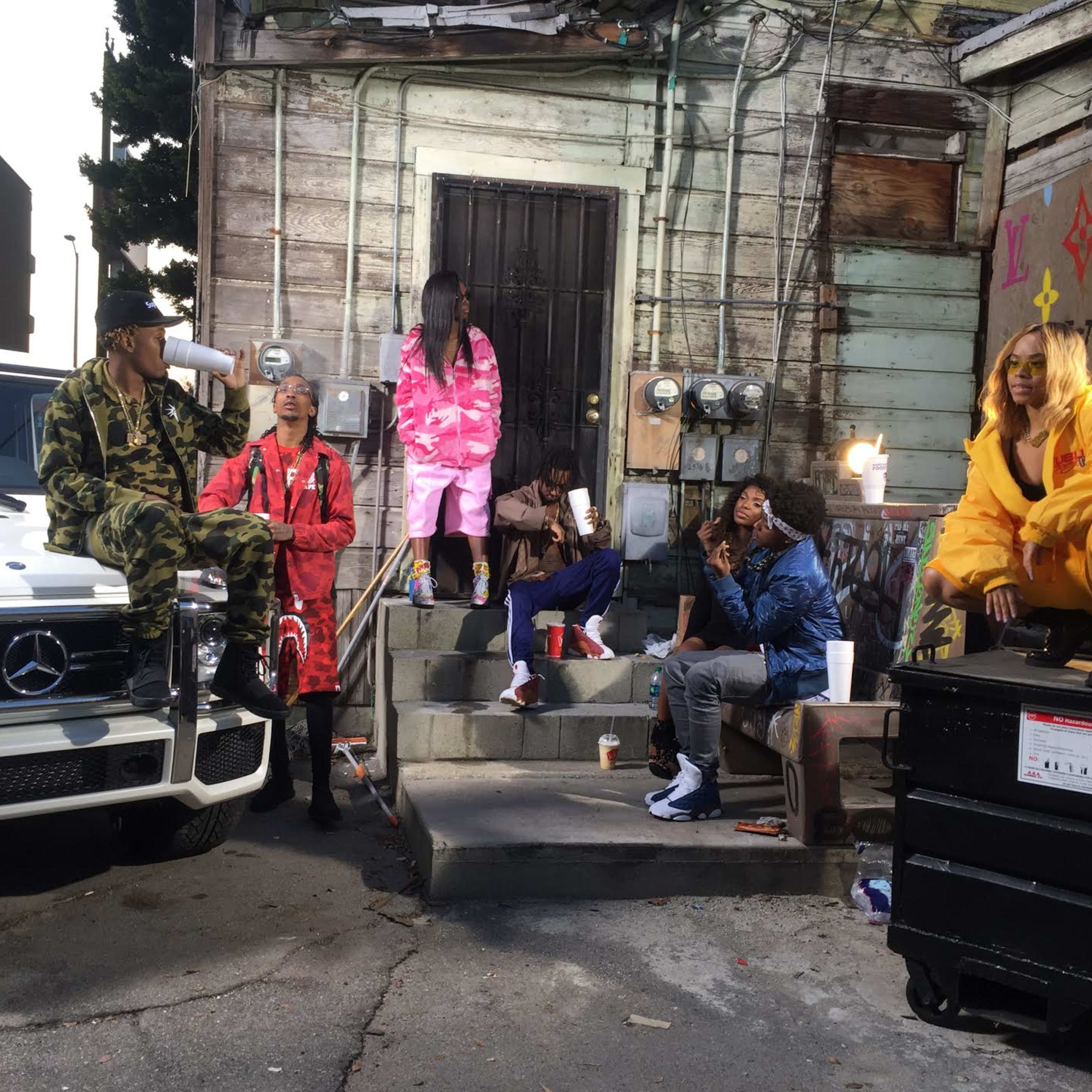 A group of people in casual clothing are gathered near a white Mercedes-Benz SUV and a dumpster outside an old wooden building, engaged in various activities.