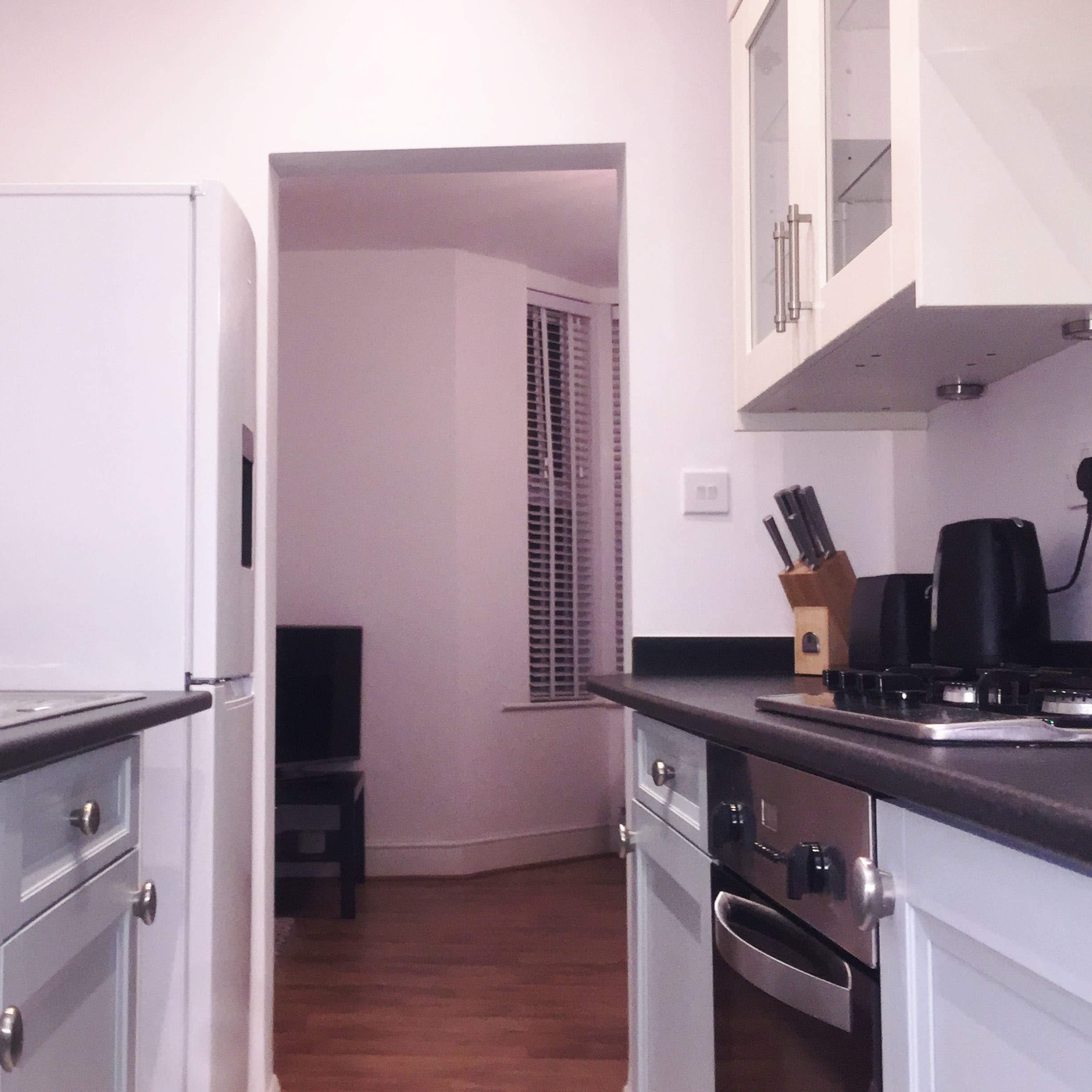 A modern kitchen features white cabinetry, a stainless steel refrigerator, and a view into a living area with a black chair.