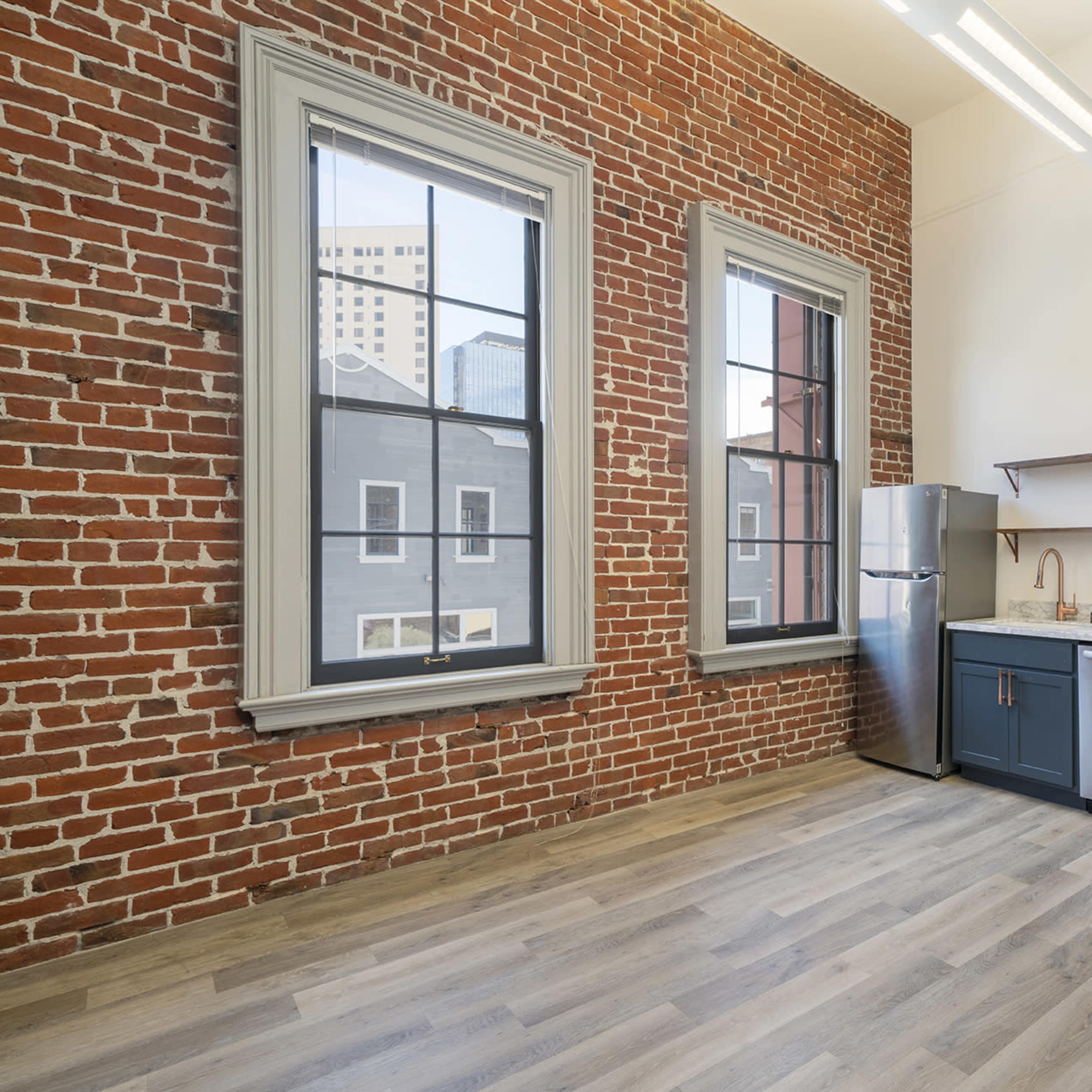 A modern kitchen area in a room with exposed brick walls and large windows, featuring a refrigerator, countertop, and open shelving.