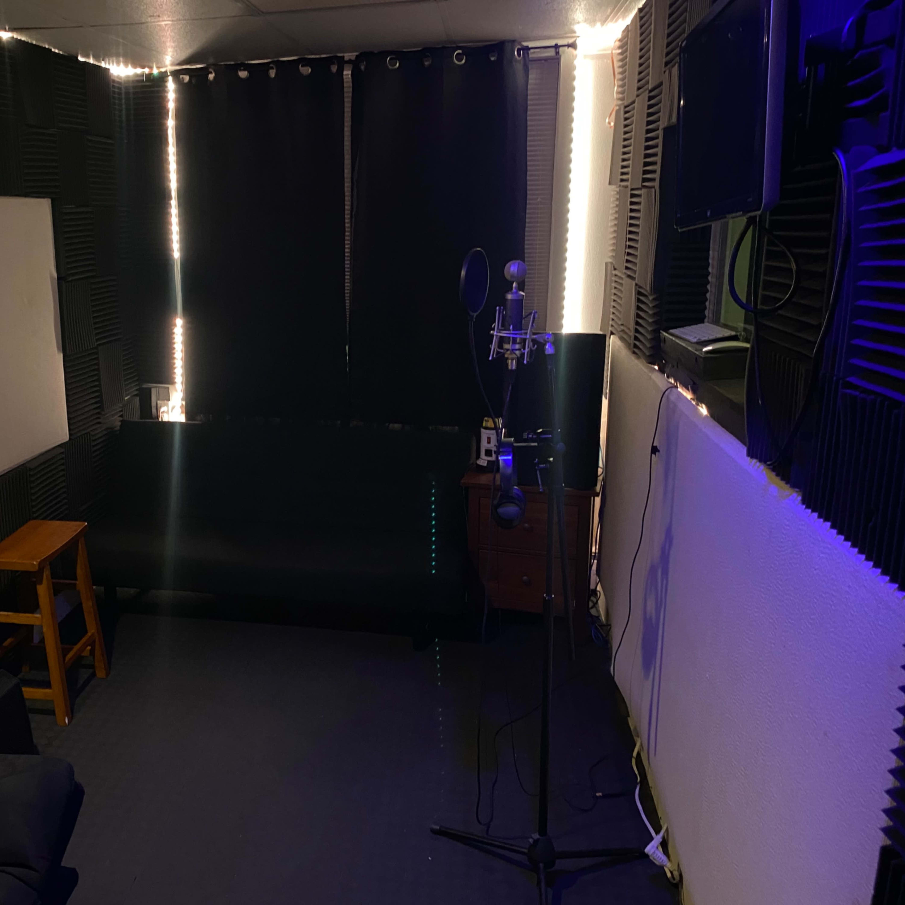 A small recording studio with soundproofing panels, a microphone on a stand, a couch, and a stool against dark curtains.