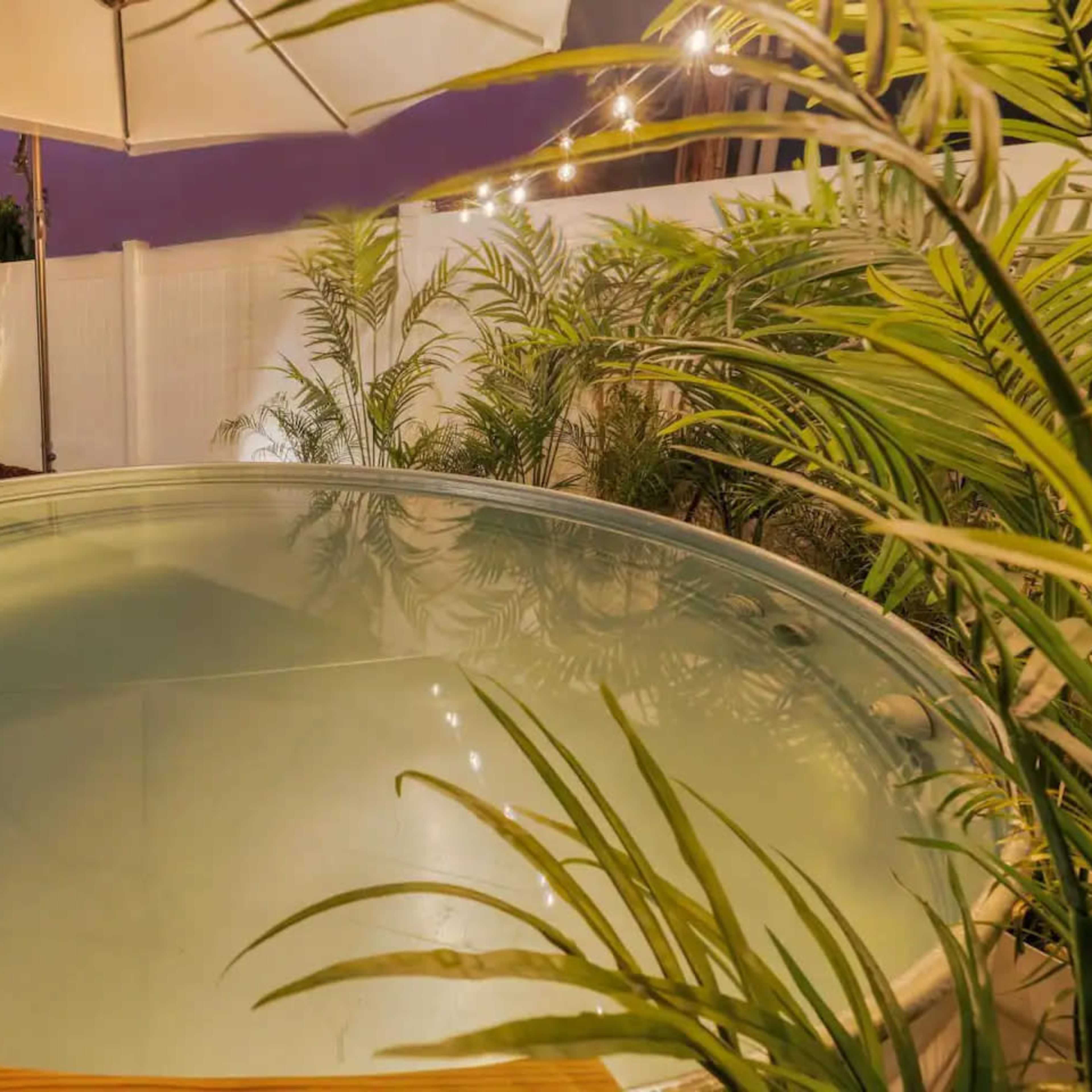 A round hot tub is set within a backyard surrounded by lush greenery and a white fence, illuminated by soft outdoor lighting.