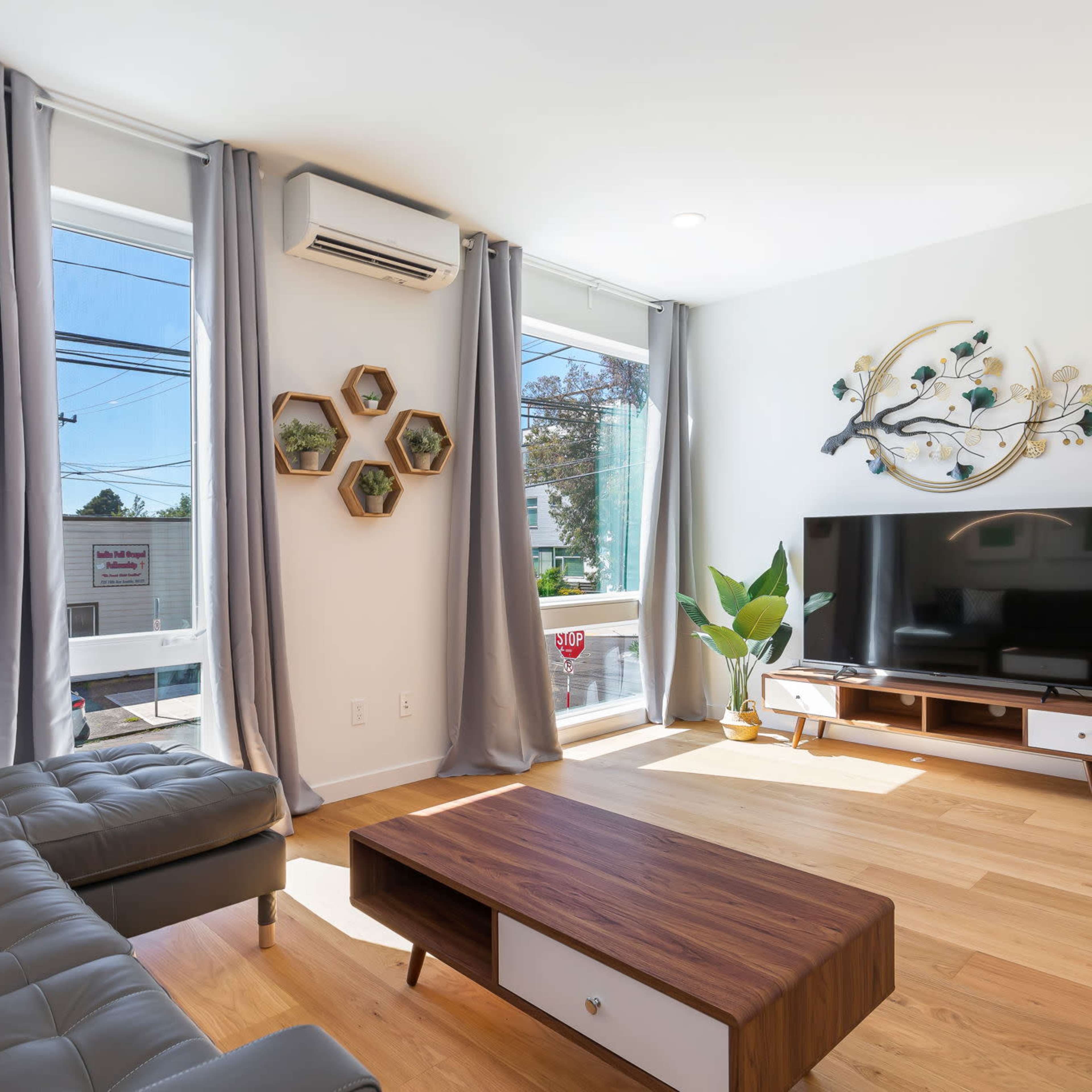 A modern living room features a gray sectional sofa, a wooden coffee table, a wall-mounted TV, and large windows with decorative plants.