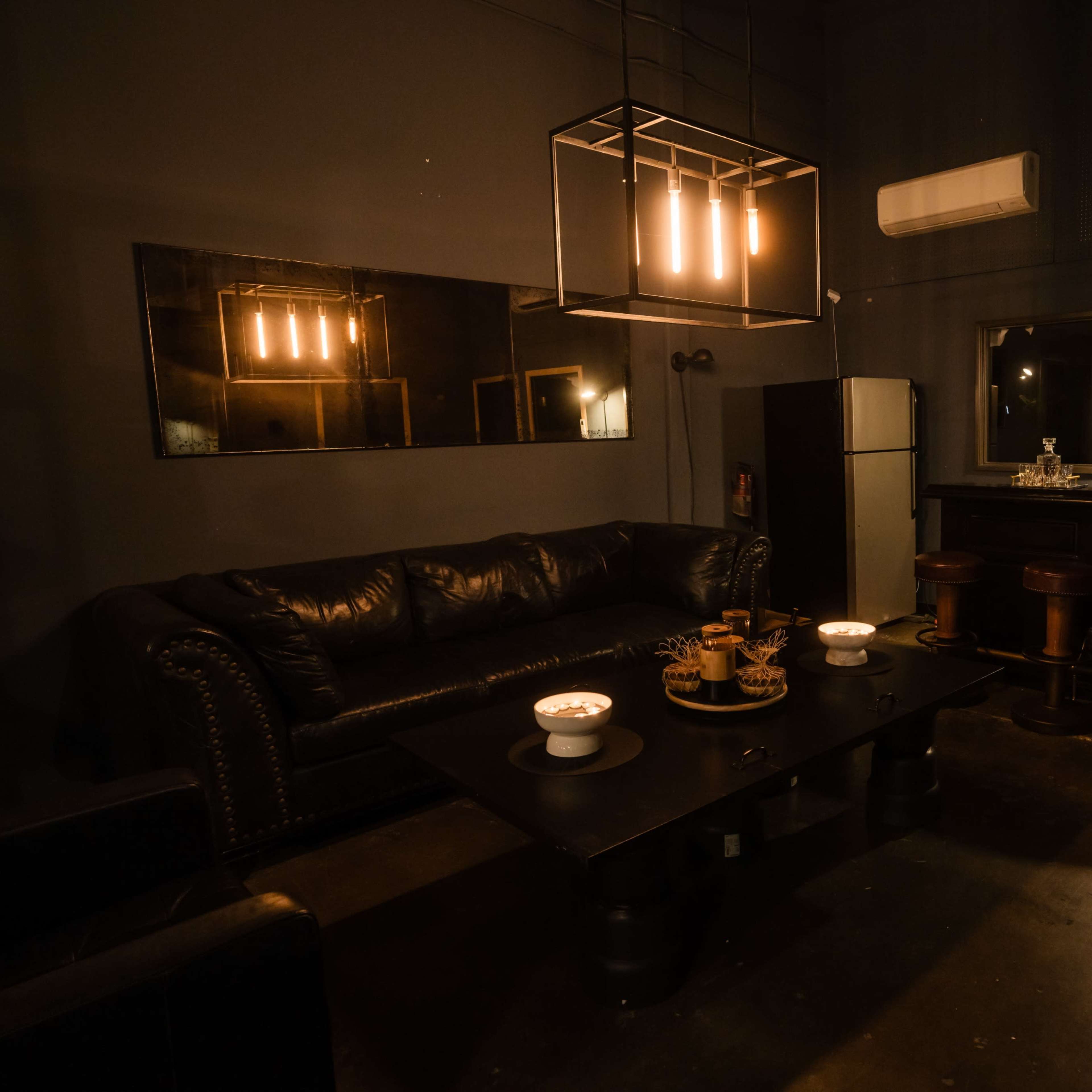 A dimly lit lounge area with a dark leather sofa, a coffee table set with candles, and a kitchenette in the background.