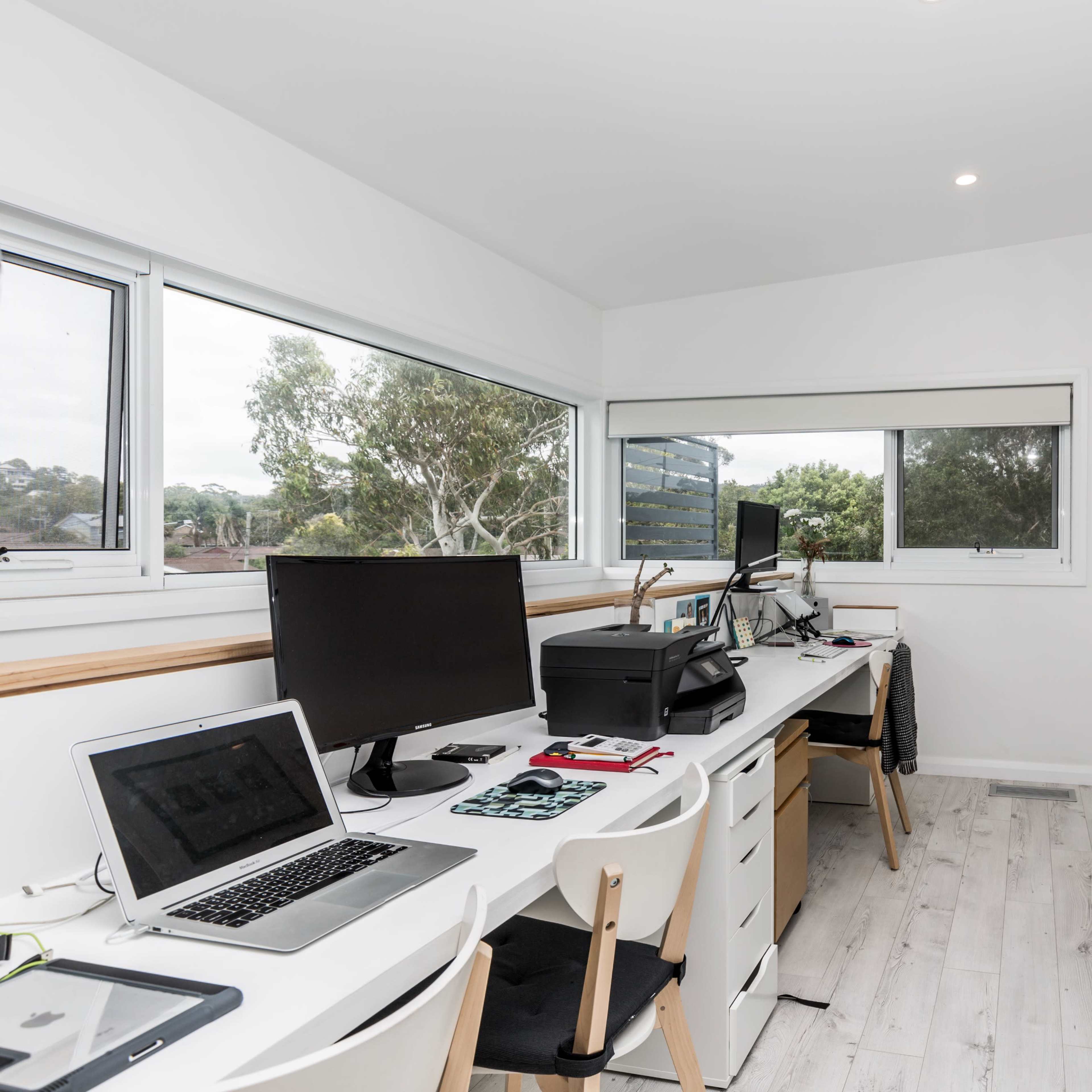 A bright home office features two desks with computers, framed photographs on the walls, and large windows offering a view of greenery.